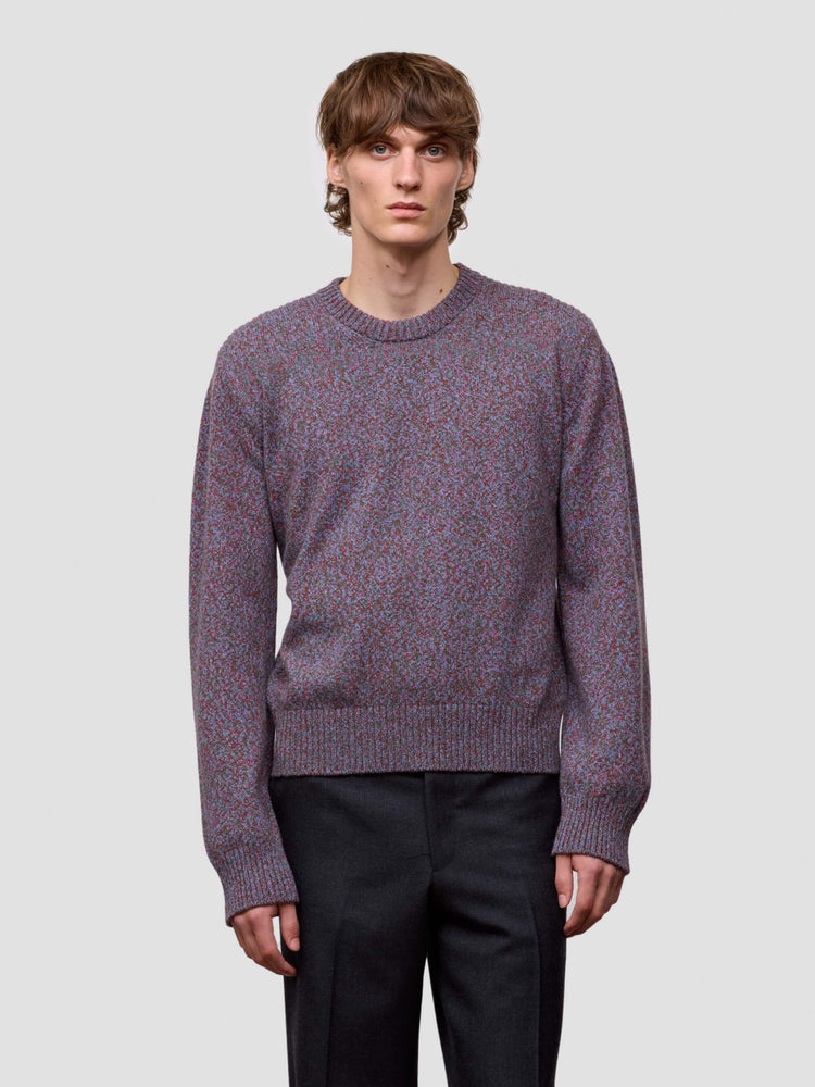 Man wearing a patterned purple cashmere sweater and black pants on a plain background