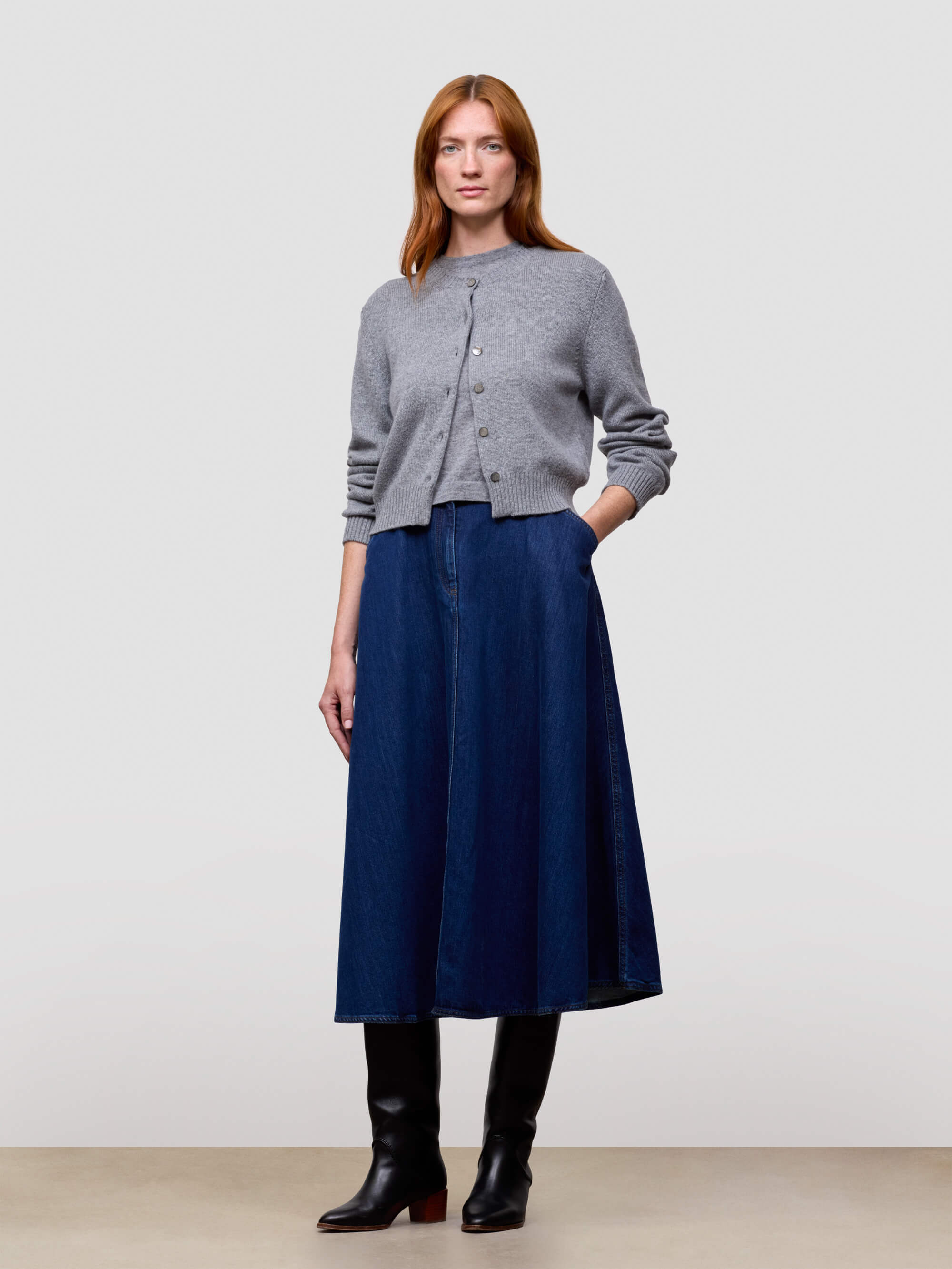 Woman wearing a gray cashmere cardigan and blue skirt and black boots on a light gray background