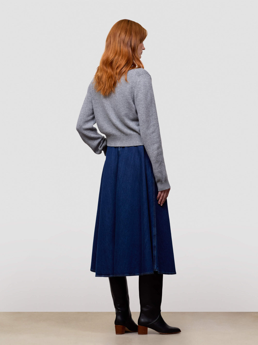 Woman wearing a gray cashmere cardigan and blue skirt and black boots on a light gray background