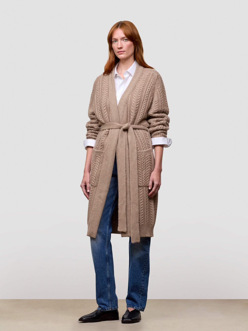 Woman wearing a long beige cashmere cardigan over a white shirt and blue jeans on a plain background