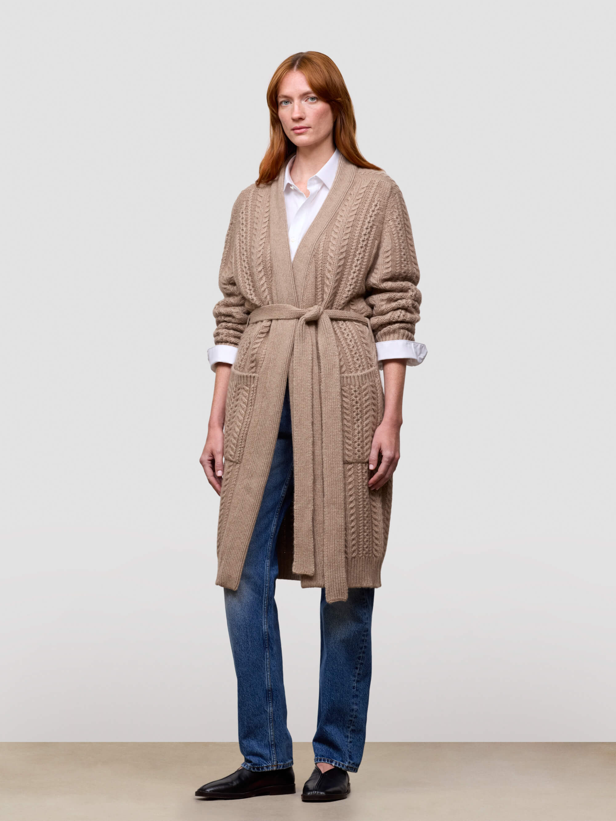 Woman wearing a long beige cashmere cardigan over a white shirt and blue jeans on a plain background