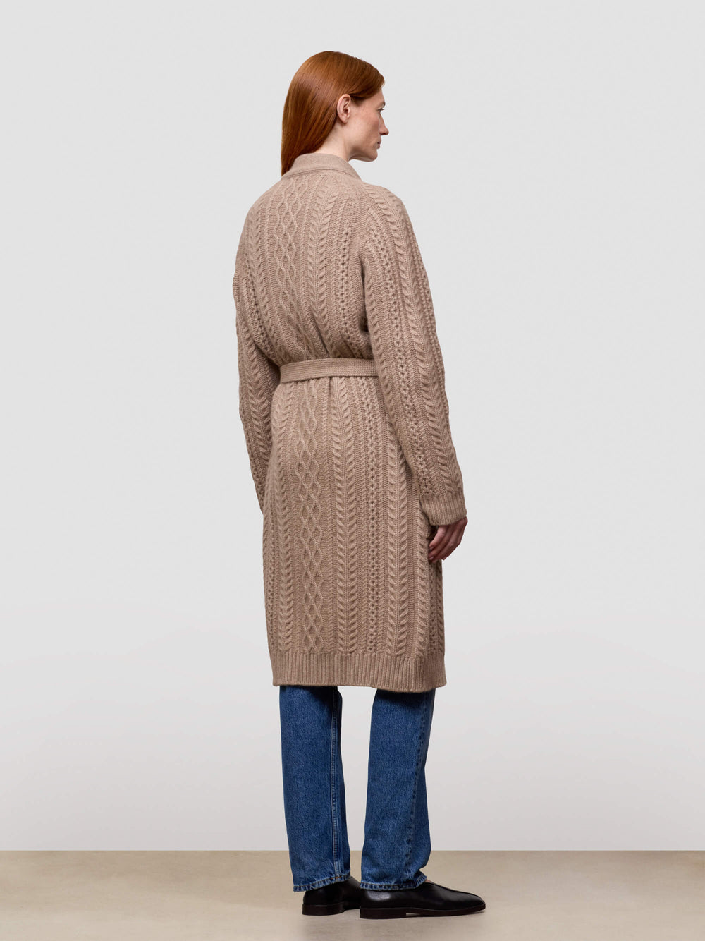 Woman wearing a long beige cable knit cashmere cardigan on a plain background