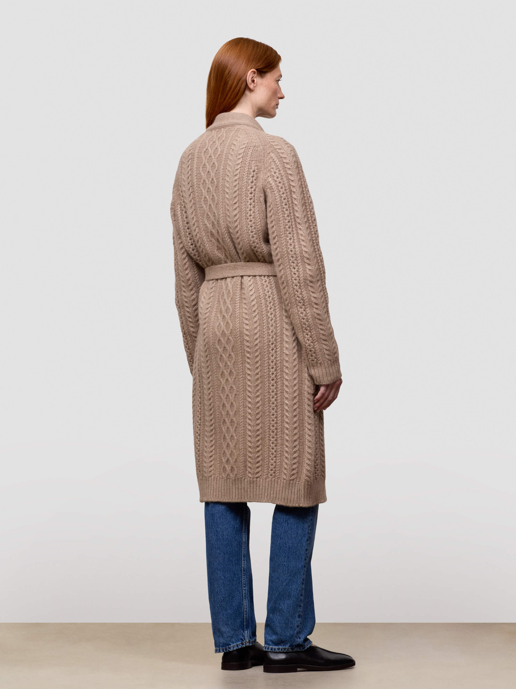 Woman wearing a long beige cable knit cashmere cardigan on a plain background