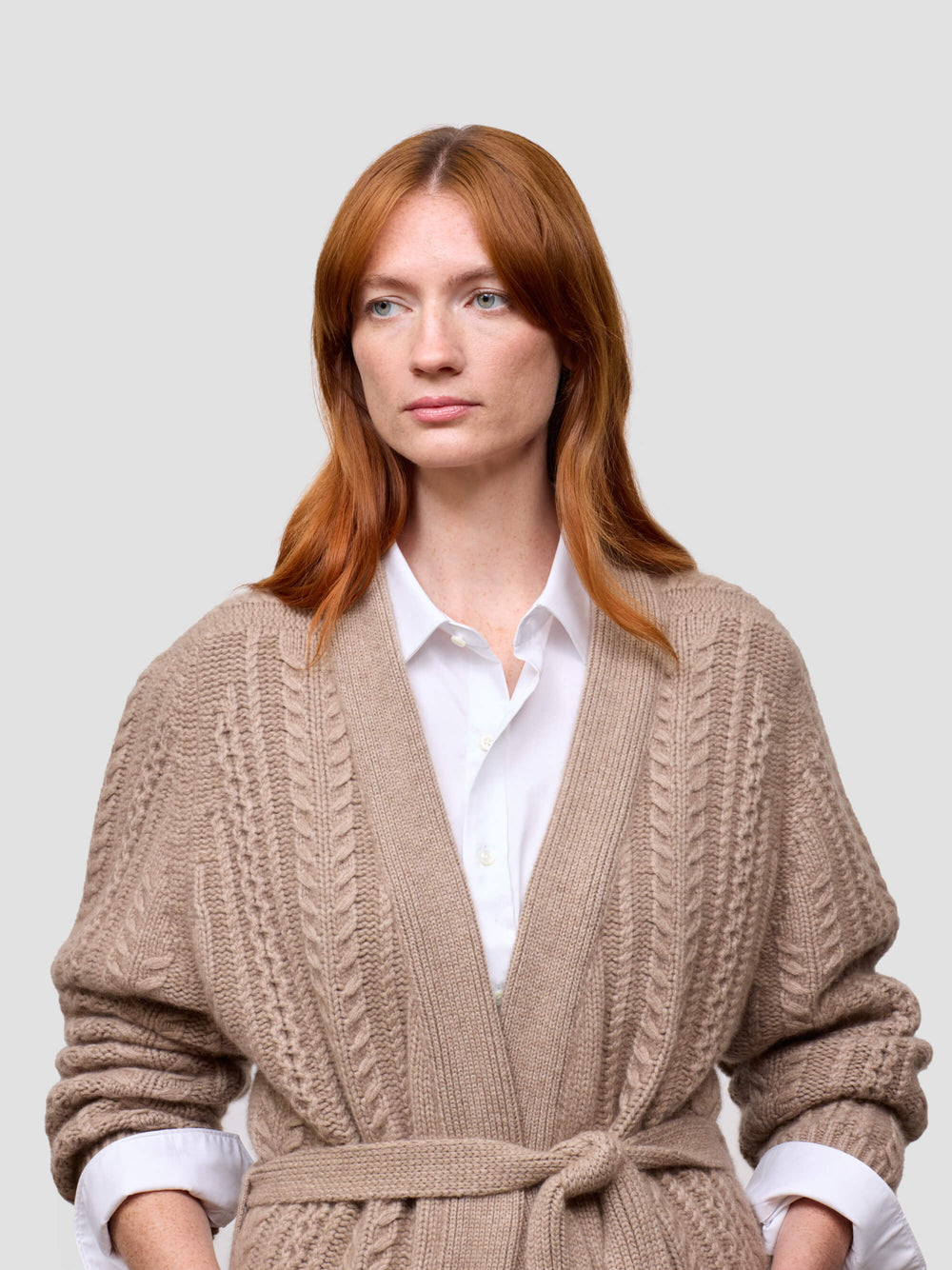 Woman wearing a beige knitted cashmere cardigan over a white shirt on a light gray background