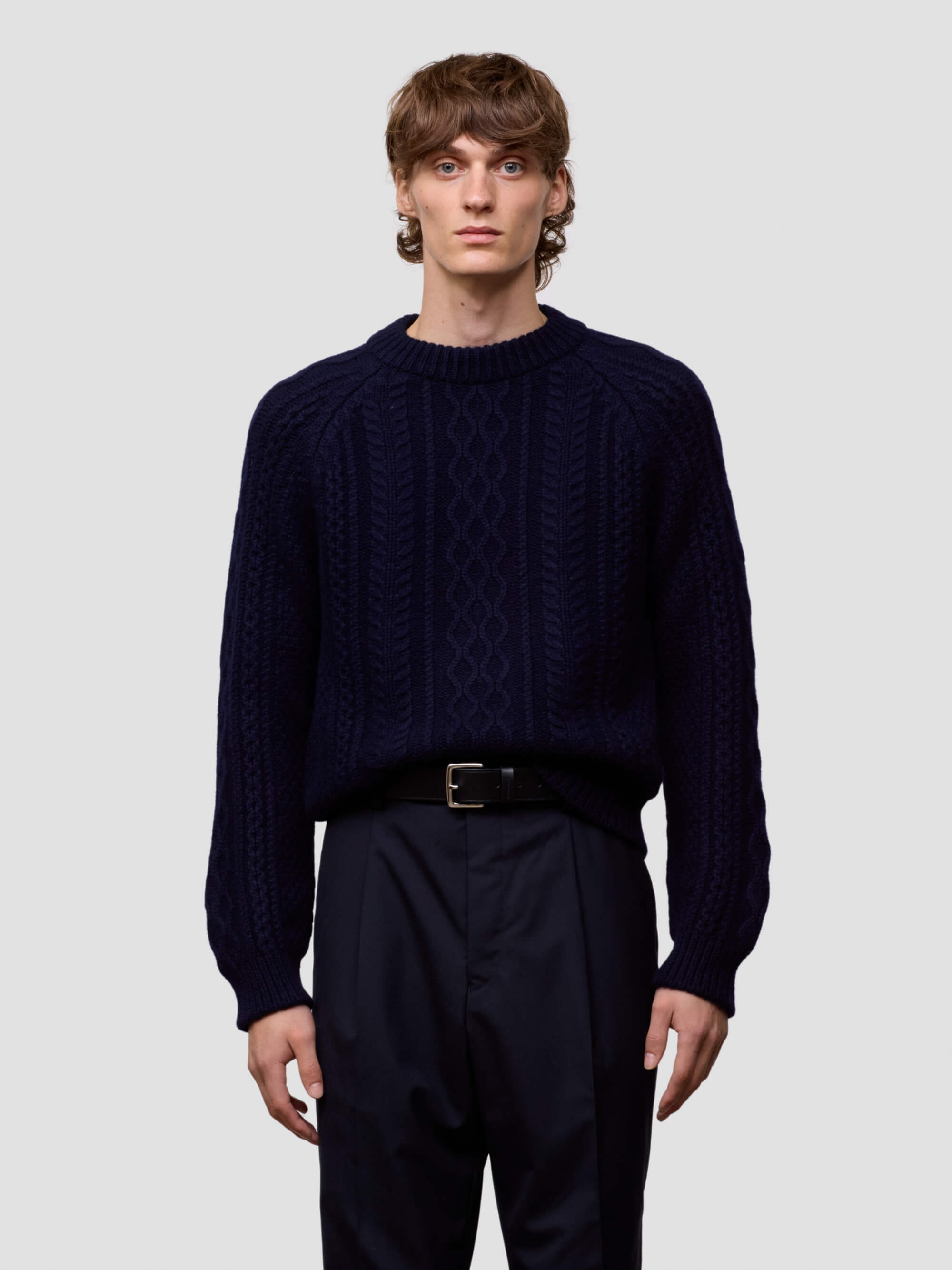 Man wearing a dark blue cable knit cashmere sweater and black pants on a light gray background