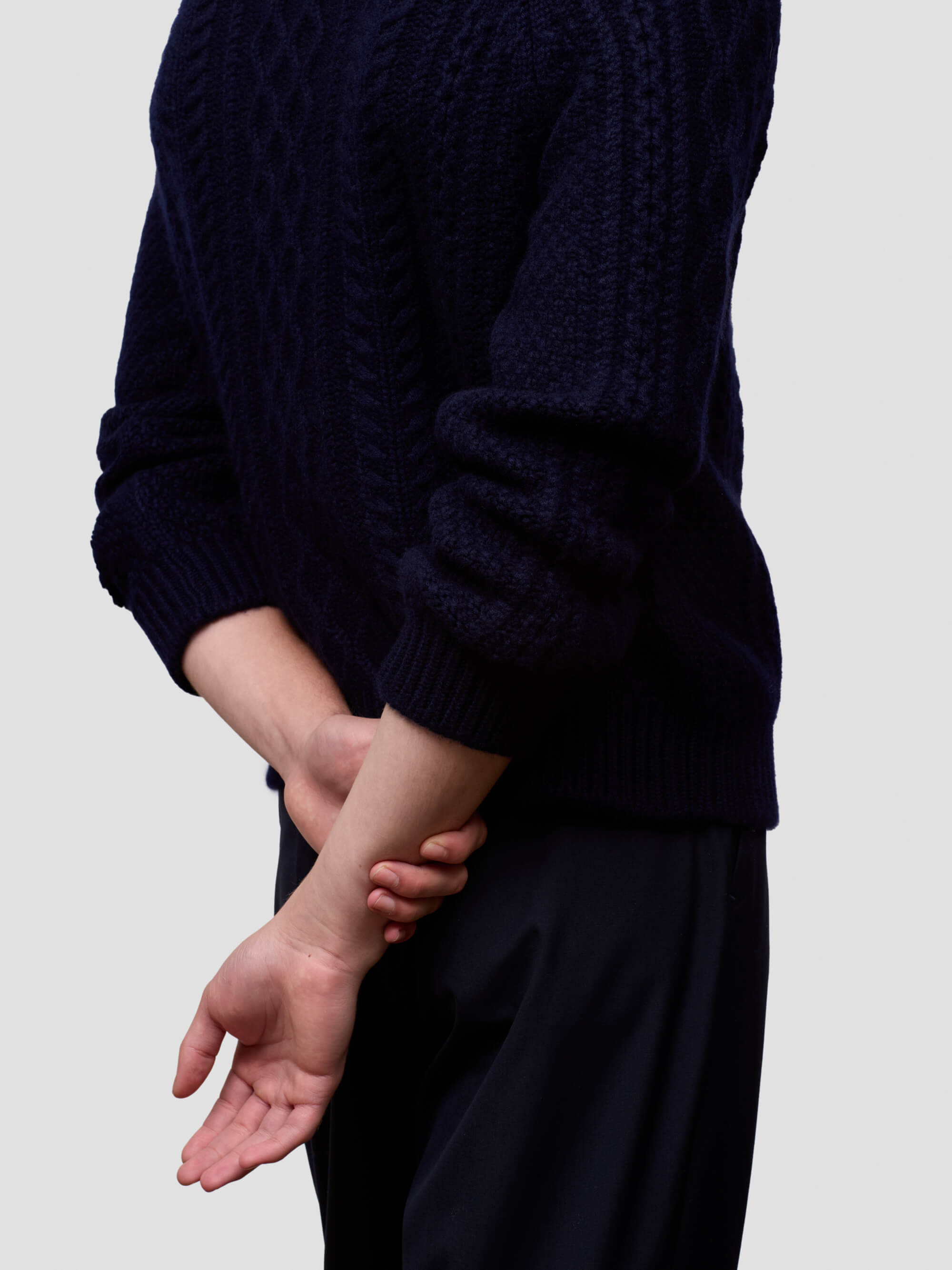 Man wearing a dark blue cable knit cashmere sweater and black pants on a light gray background