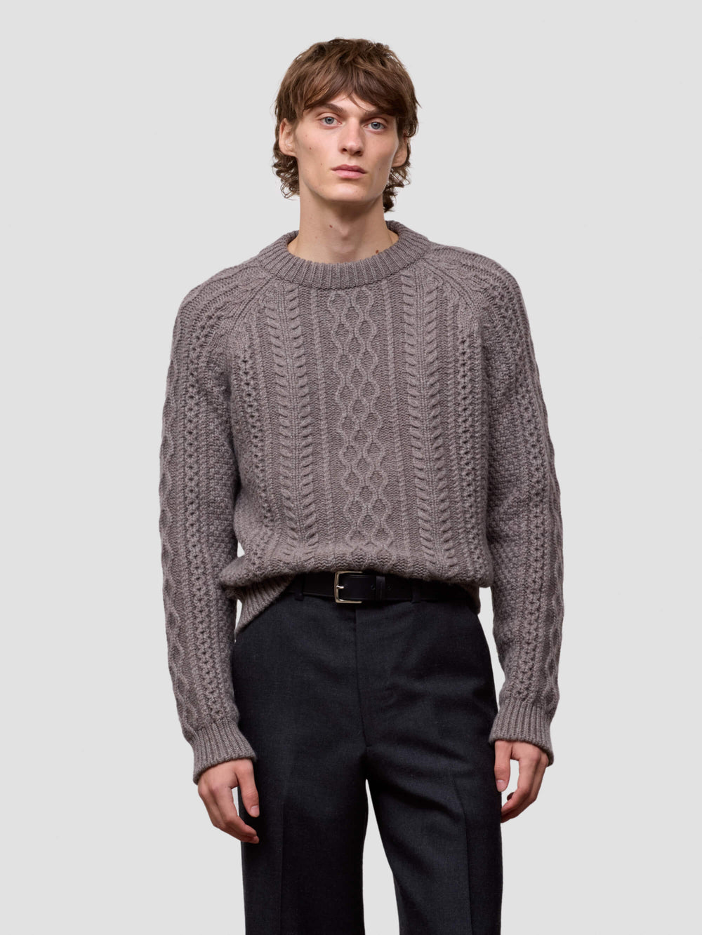 Man wearing a brown cable knit cashmere sweater and black pants on a light gray background