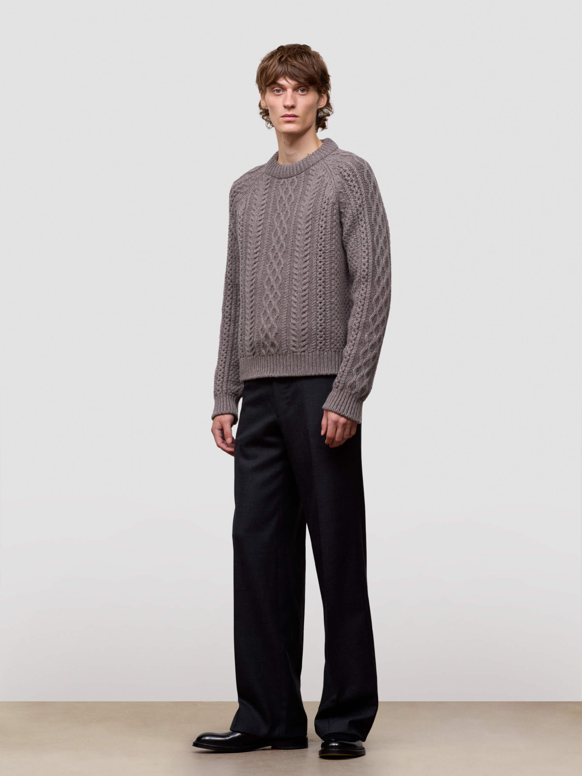 Man wearing a brown cable knit cashmere sweater and black pants on a light gray background