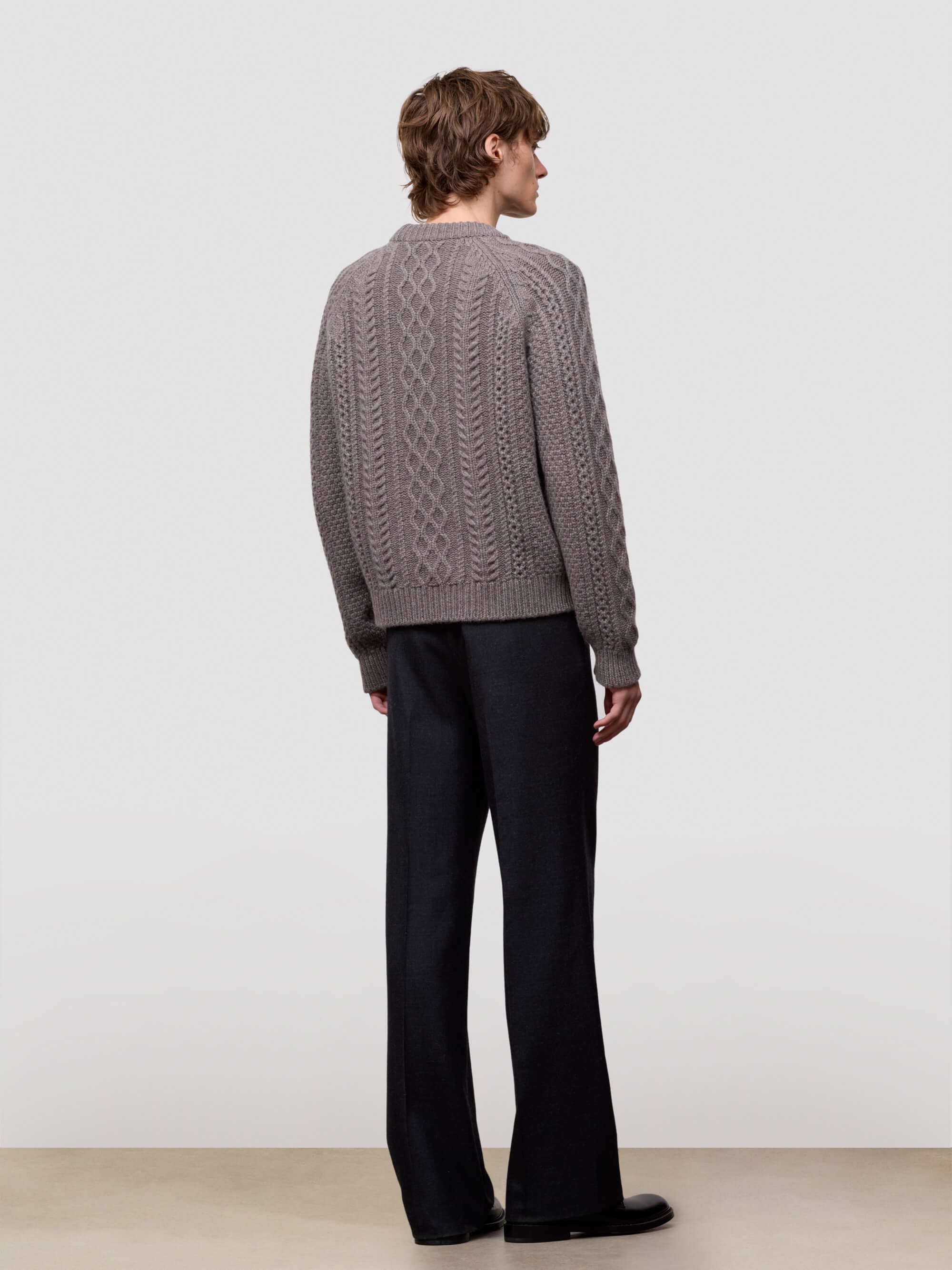 Man wearing a brown cable knit cashmere sweater and black pants on a light gray background
