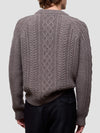 Man wearing a brown cable knit cashmere sweater and black pants on a light gray background