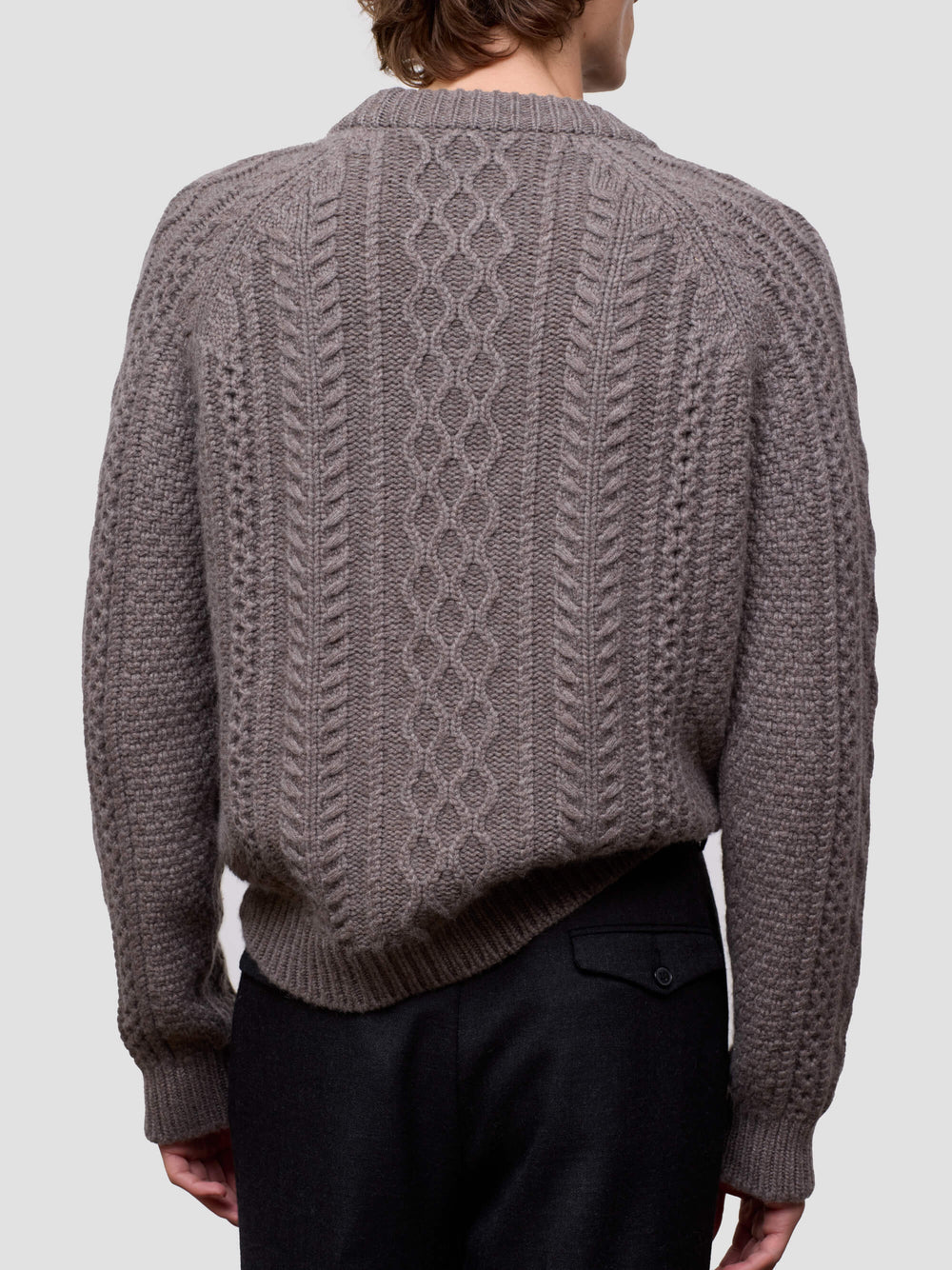 Man wearing a brown cable knit cashmere sweater and black pants on a light gray background