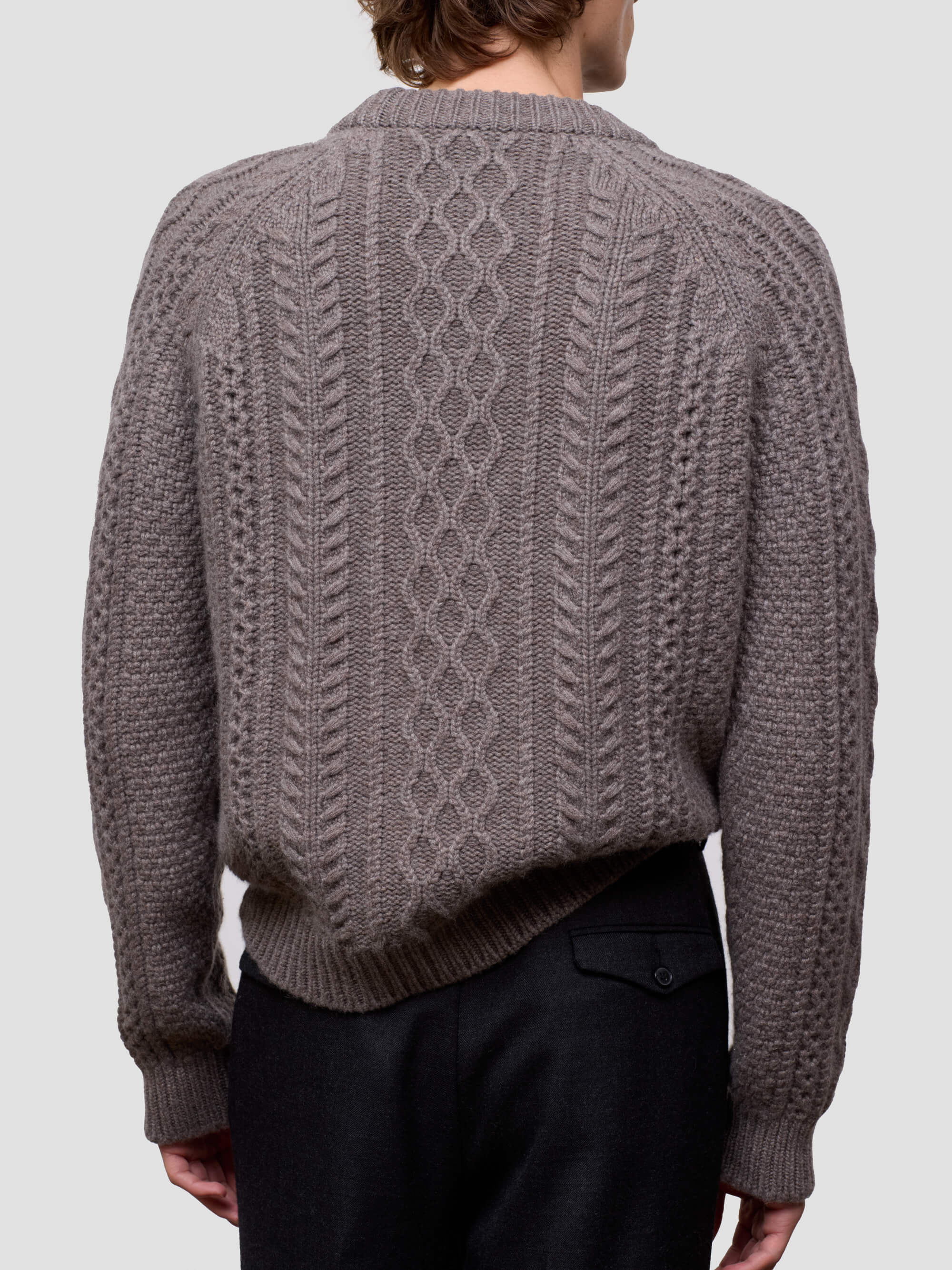 Man wearing a brown cable knit cashmere sweater and black pants on a light gray background