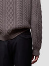 Man wearing a brown cable knit cashmere sweater and black pants on a light gray background