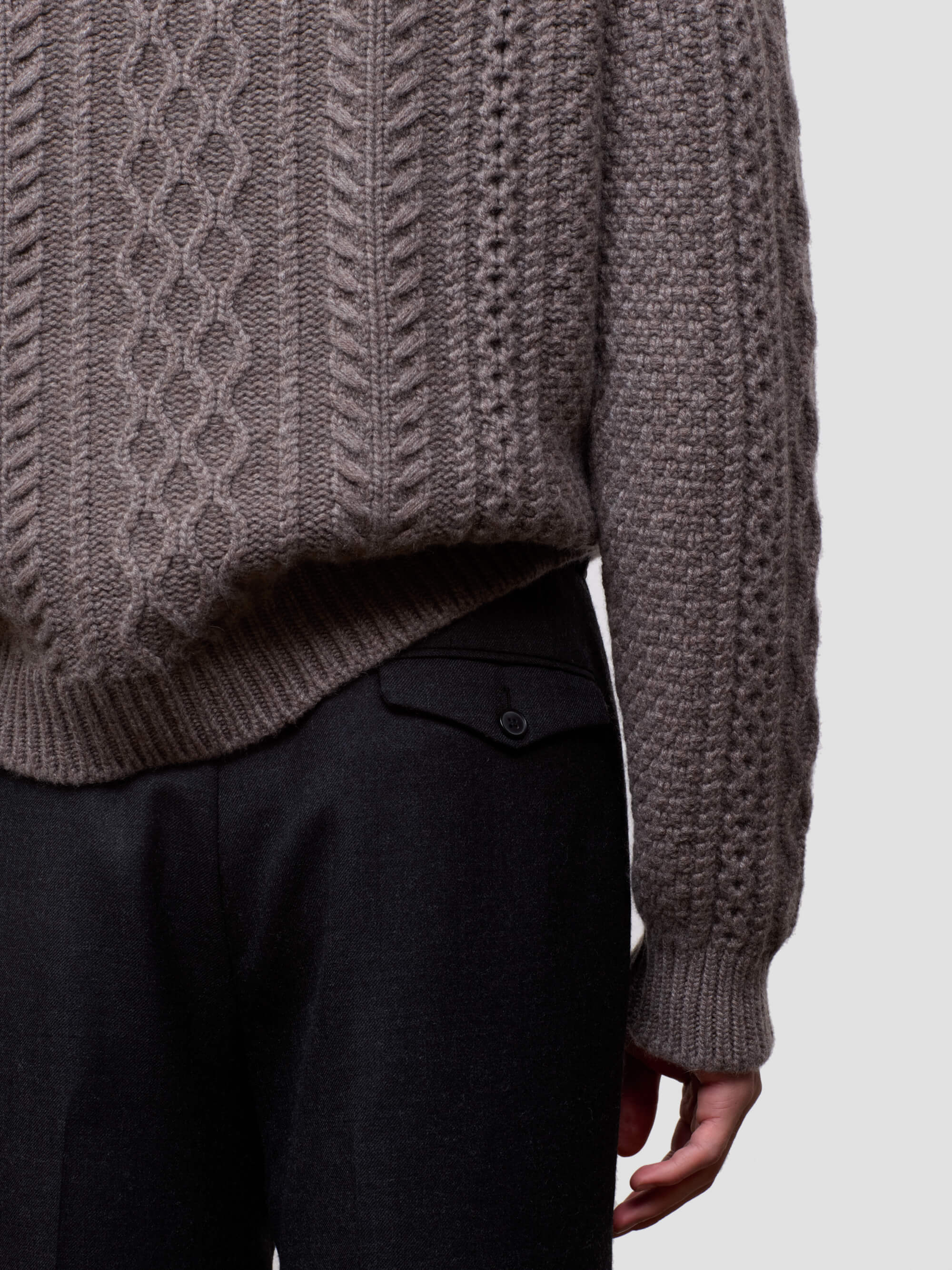 Man wearing a brown cable knit cashmere sweater and black pants on a light gray background