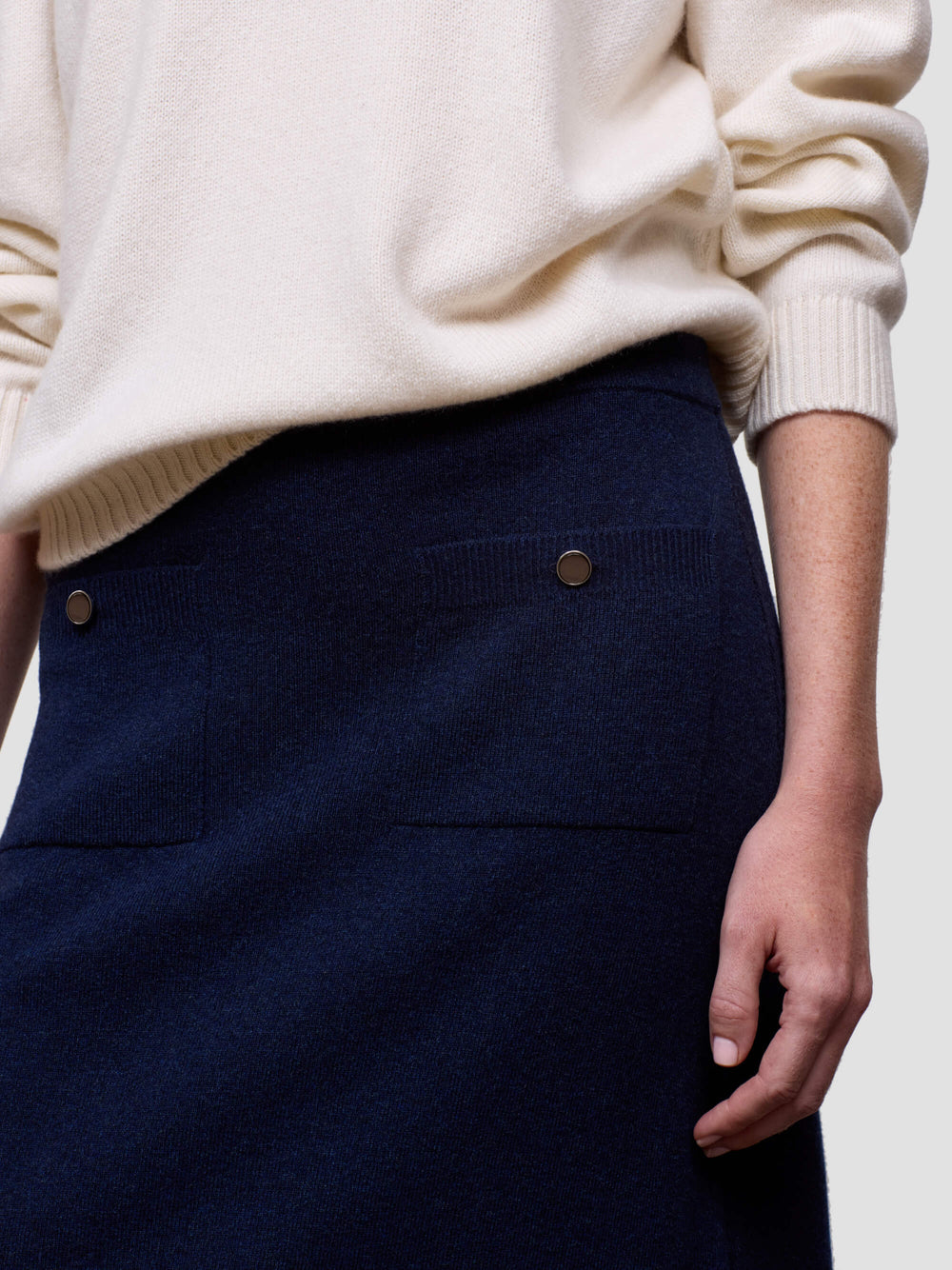 Woman wearing a white cashmere sweater and navy cashmere skirt against a plain background