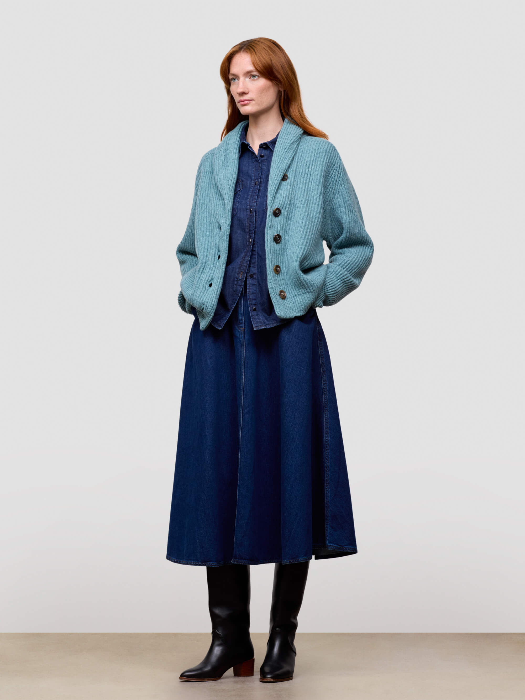 Woman wearing a teal knitted cashmere cardigan over a blue dress and black boots on a light gray background