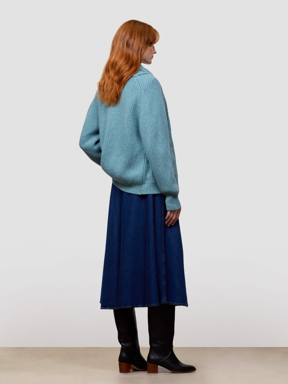 Woman wearing a teal knitted cashmere cardigan over a blue dress and black boots on a light gray background