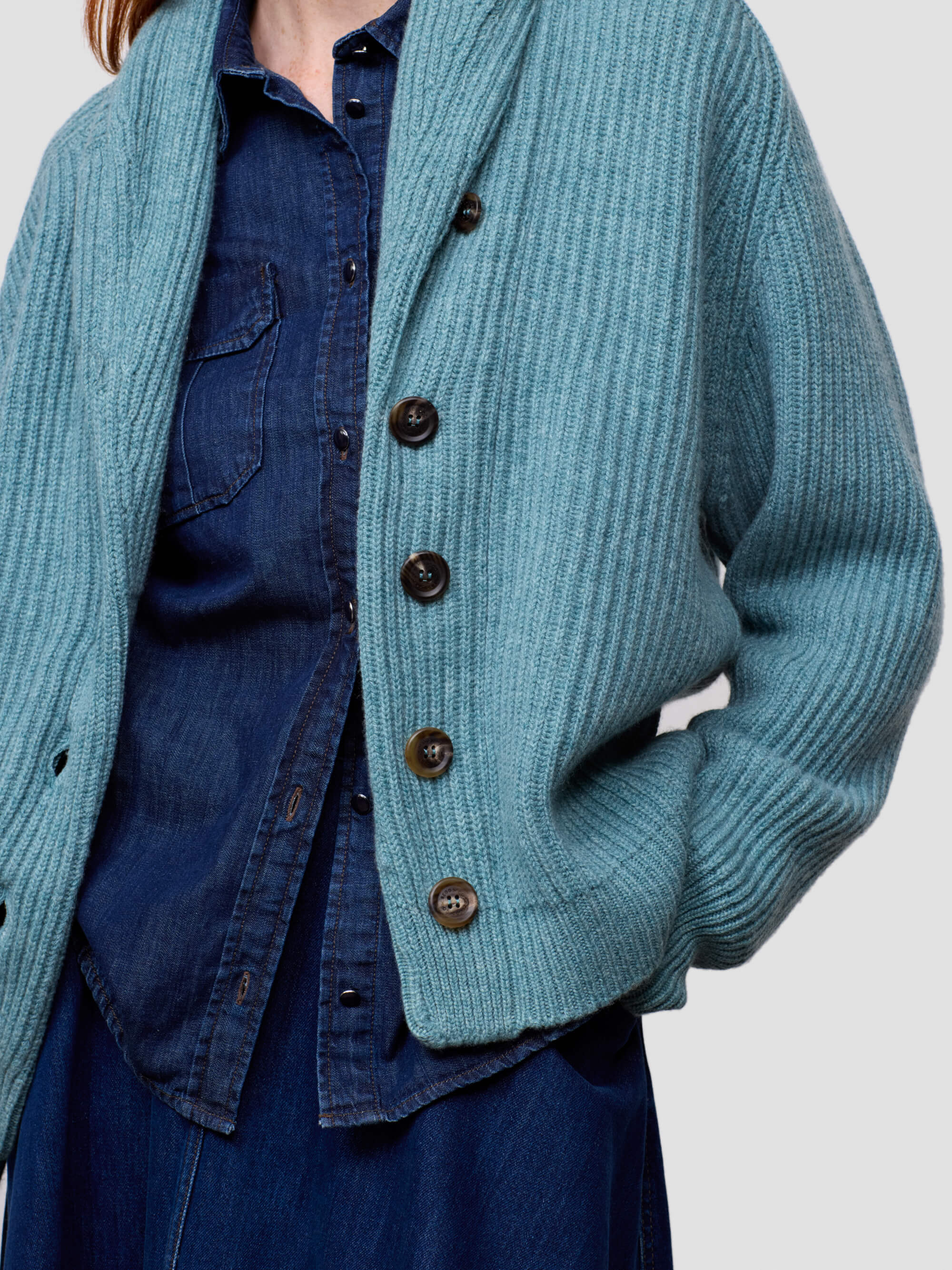 woman wearing a teal ribbed cashmere cardigan over a blue denim shirt on a plain background