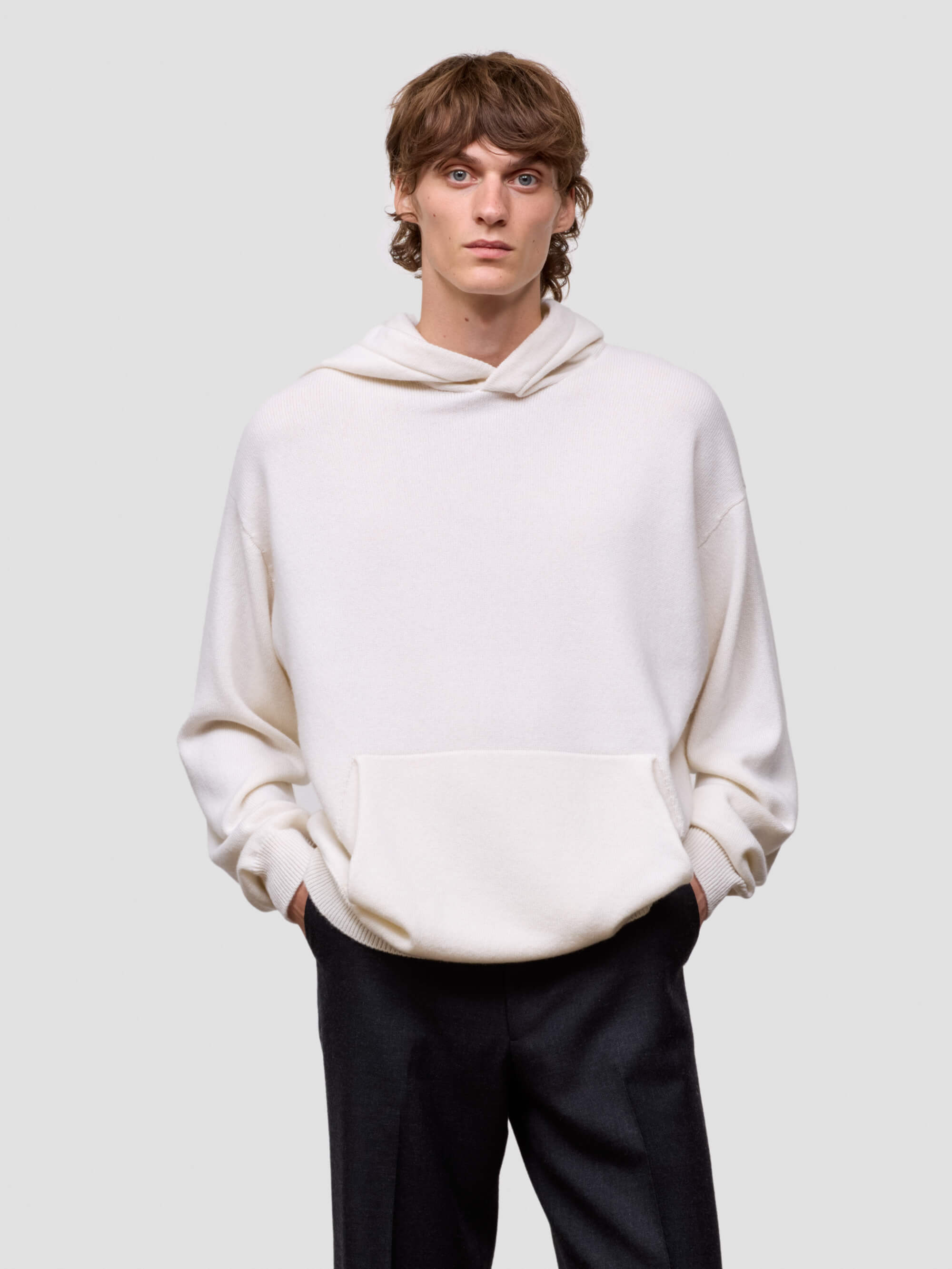 man wearing a white cashmere hoodie and black pants on a light gray background