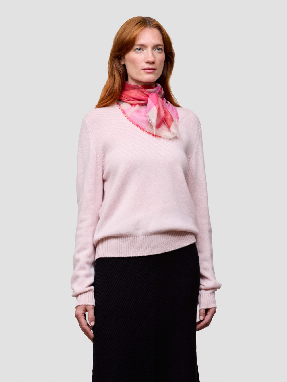 Woman wearing a light pink sweater and black skirt with a pink scarf on a white background