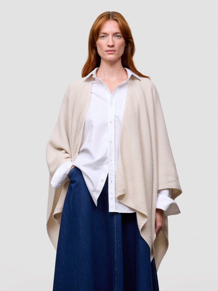 Woman wearing a beige poncho over a white shirt and blue pants on a plain background