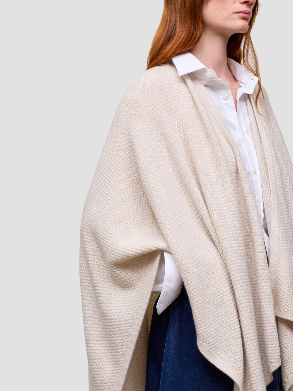 Person wearing a beige poncho over a white shirt and blue jeans on a plain background