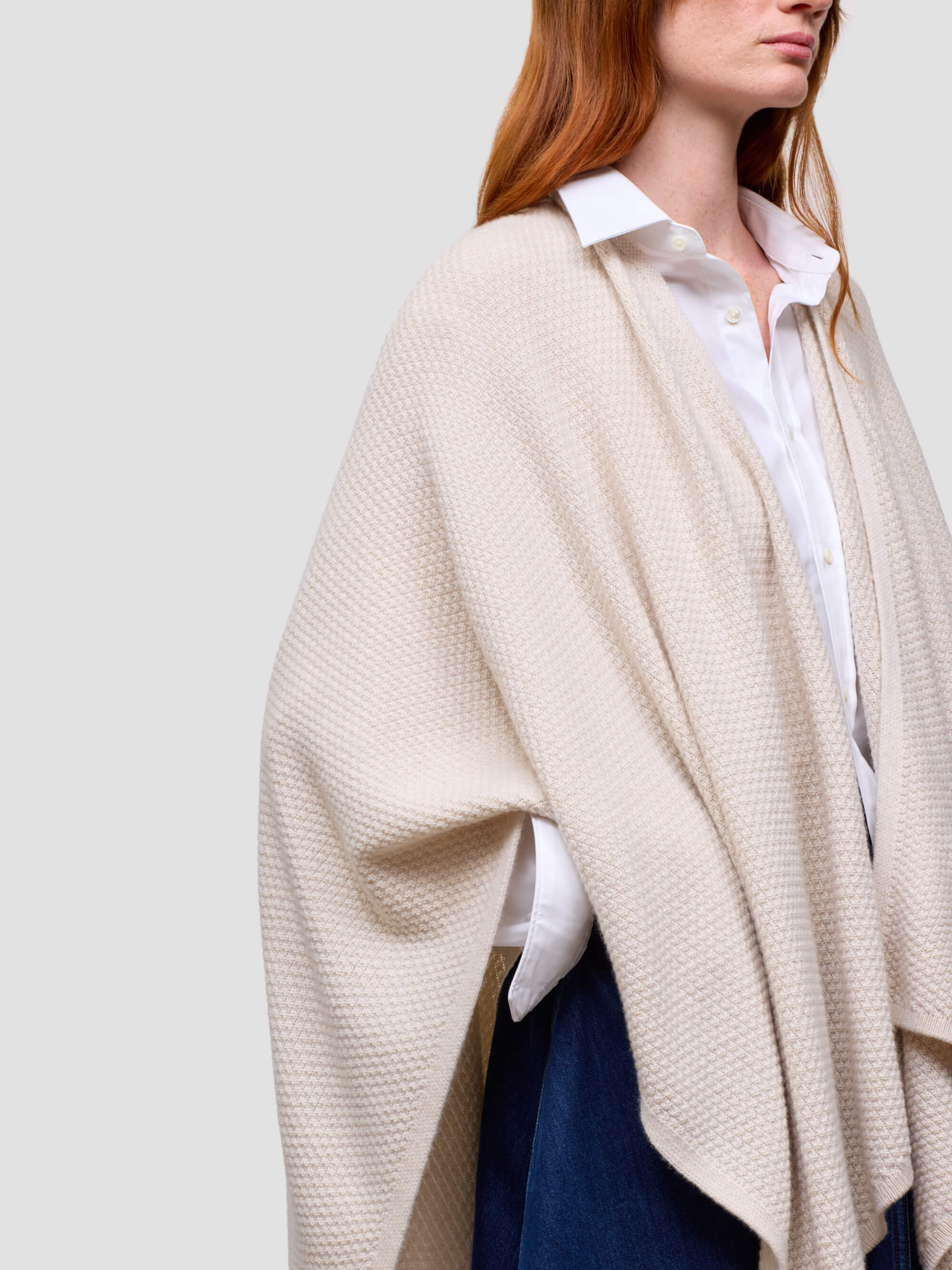 Person wearing a beige poncho over a white shirt and blue jeans on a plain background