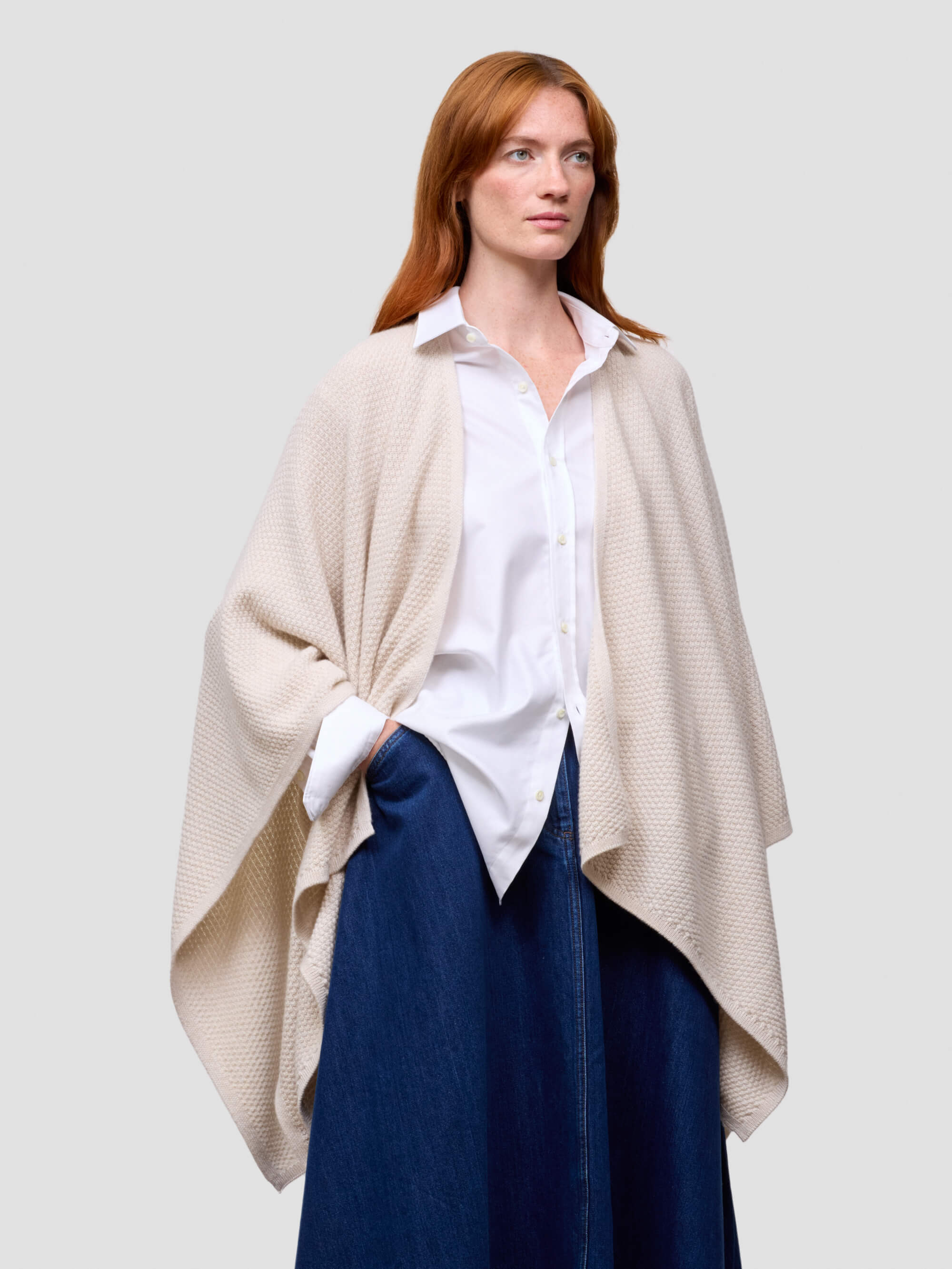 Woman wearing a beige cashmere poncho over a white shirt and blue pants on a light gray background