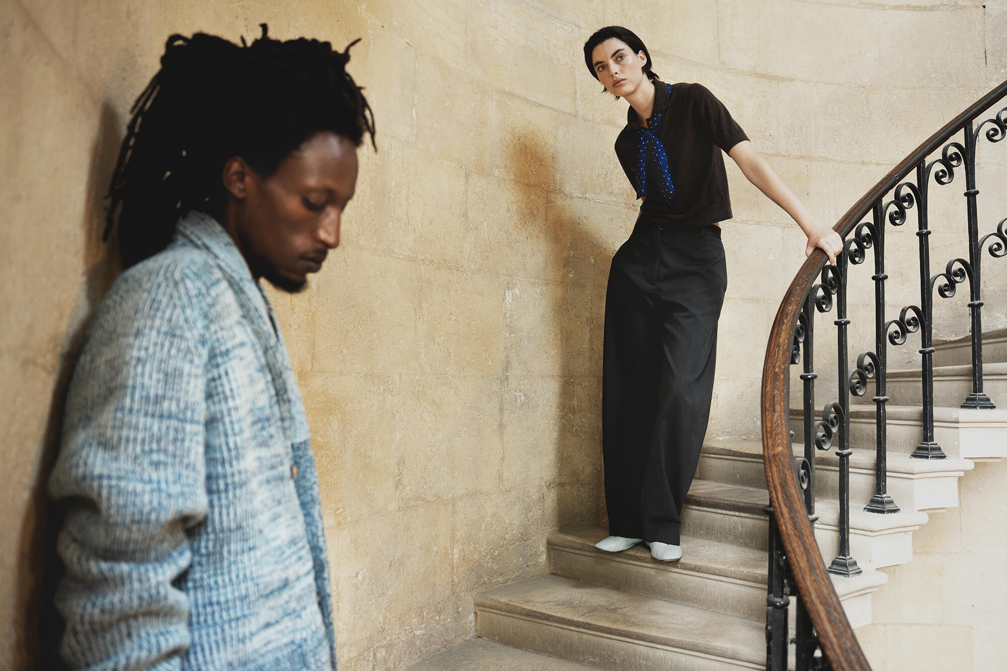 Two models in a stone stairwell setting. One model in the foreground is wearing a light blue textured knit cashmere cardigan, looking down. The other wearing a brown cashmere polo and blue polkadot scarf