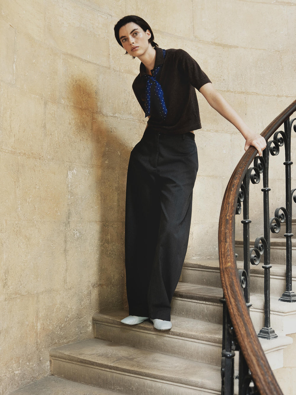 woman wearing a brown cashmere polo, black trousers and blue polkadot cashmere scarf. Standing on a stone staircase holding the railing