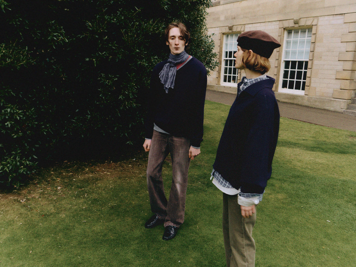 Two models standing on a green lawn in front of a stone building, wearing dark navy and brown outerwear with plaid shirts and trousers.