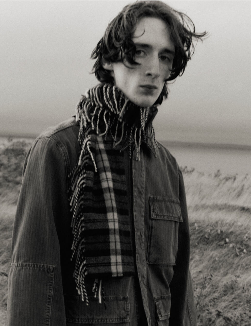 A black and white portrait of a young man outdoors wearing a dark jacket and a thick plaid scarf with fringe.