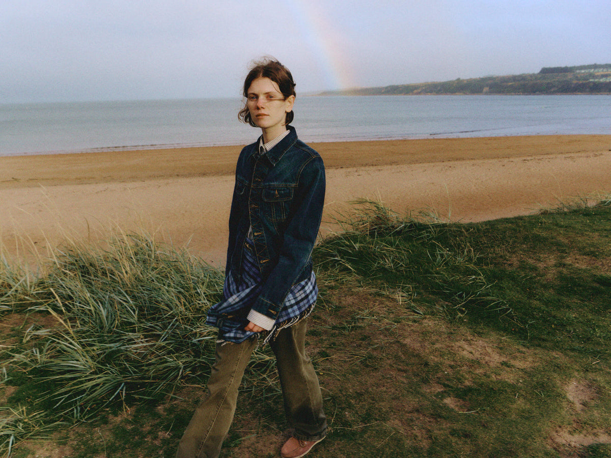 A woman standing on grassy dunes near a beach, wearing a dark denim jacket over a blue plaid scarf around her waist and olive pants, with a beach, water, and a faint rainbow in the background.