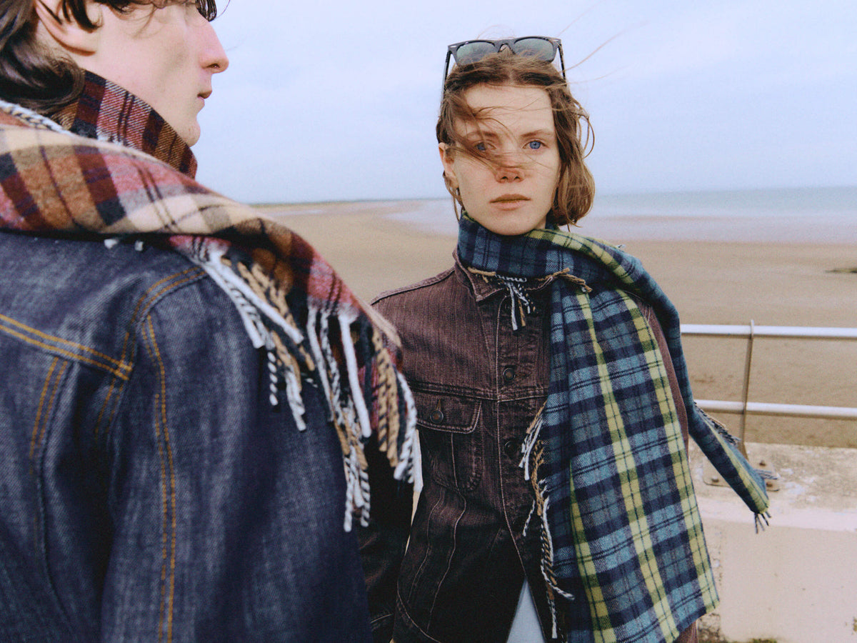 Two models standing outdoors near a beach, each wearing a dark denim jacket and a plaid scarf. The model on the right has a blue, green, and yellow plaid scarf, and the model on the left has a red, brown, and black plaid scarf with white fringe.