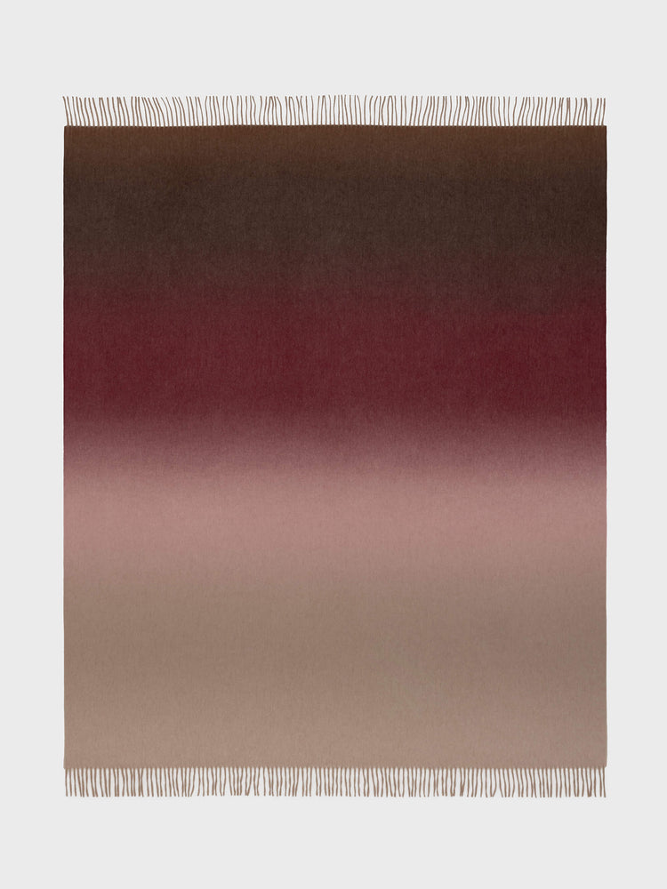 A Nuance Ombre throw blanket with a gradient of colors ranging from beige to red, featuring a subtle blend of shades with fringe detailing on the edges.
