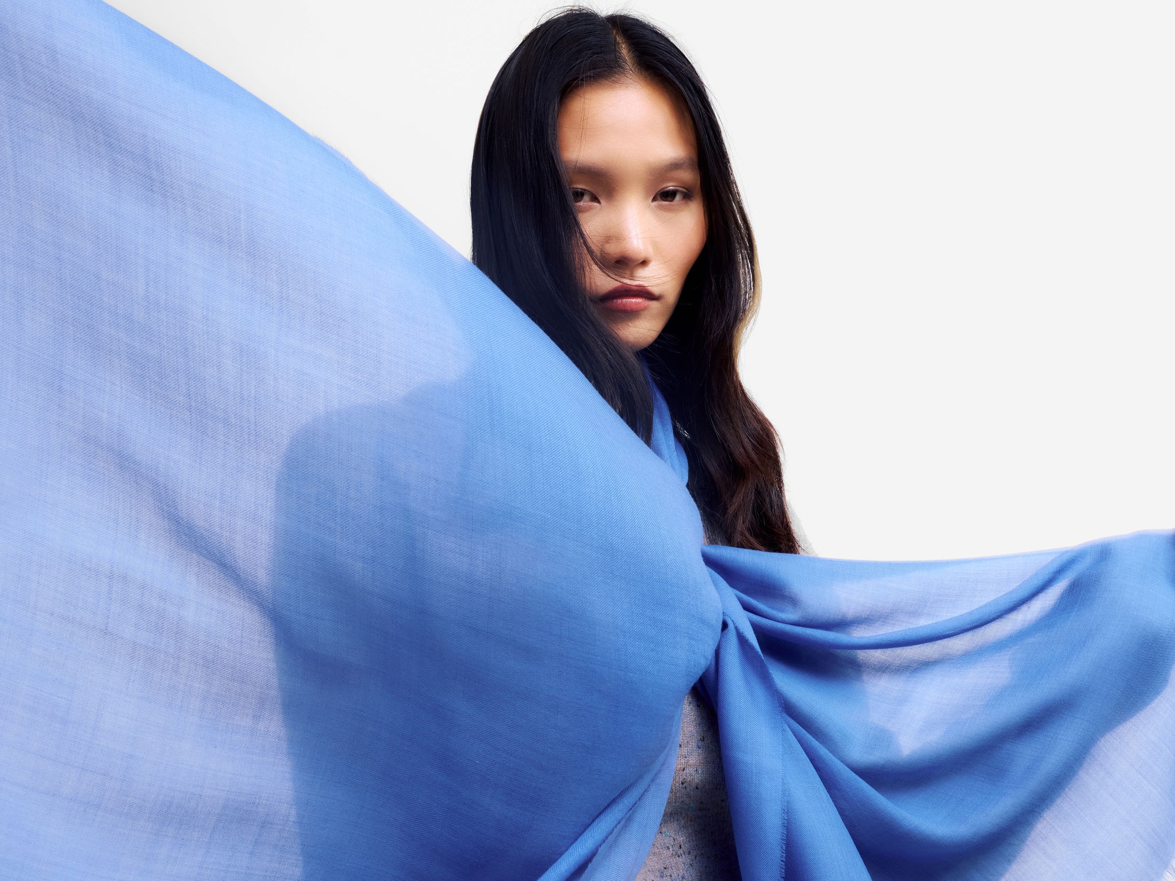 A woman with long dark hair is shown from the chest up, wrapped in a flowing sheer light blue cashmere scarf that drapes across the frame against a plain white background.