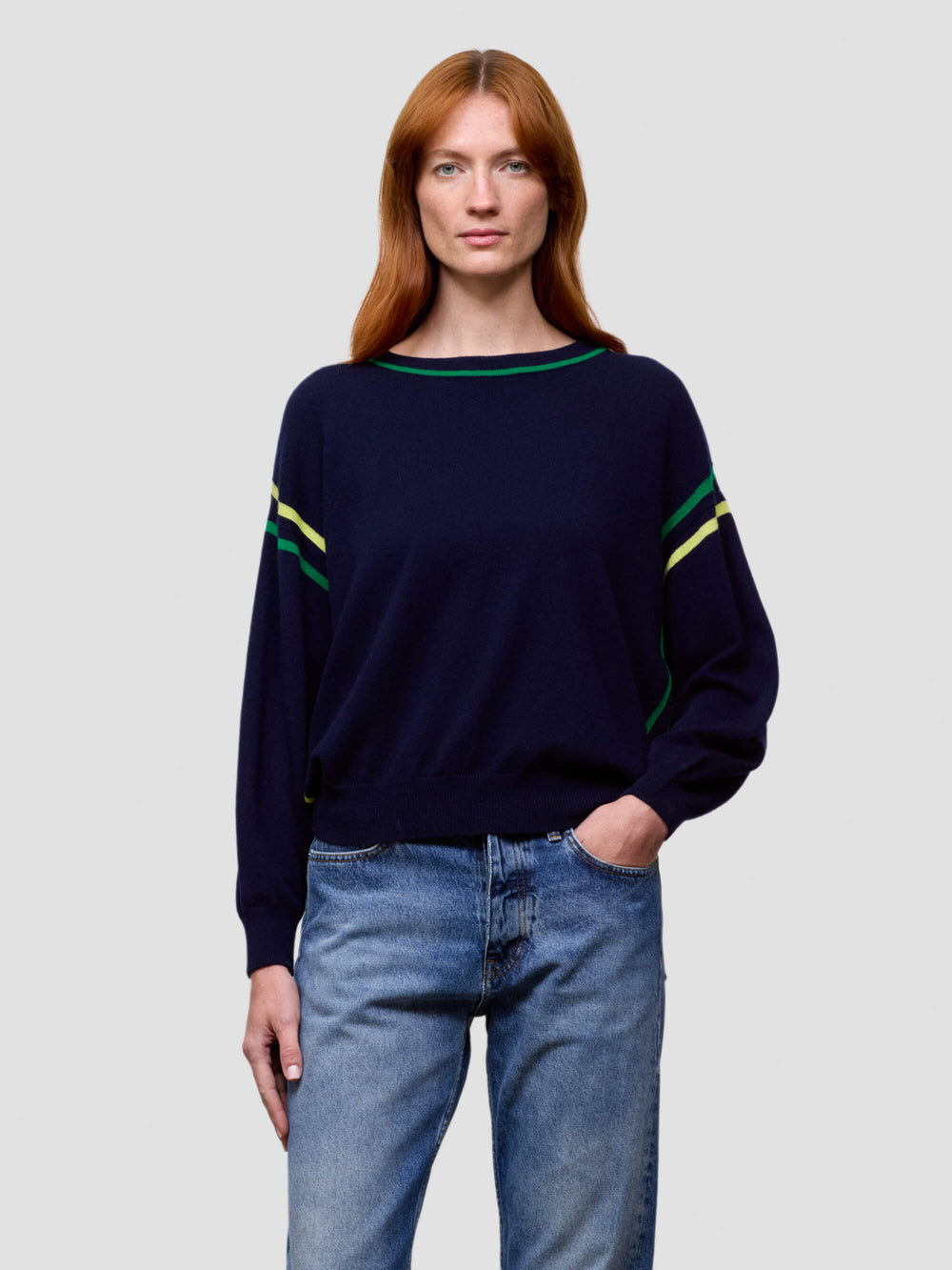 Woman wearing a navy sweater with green and yellow accents and blue jeans on a light gray background