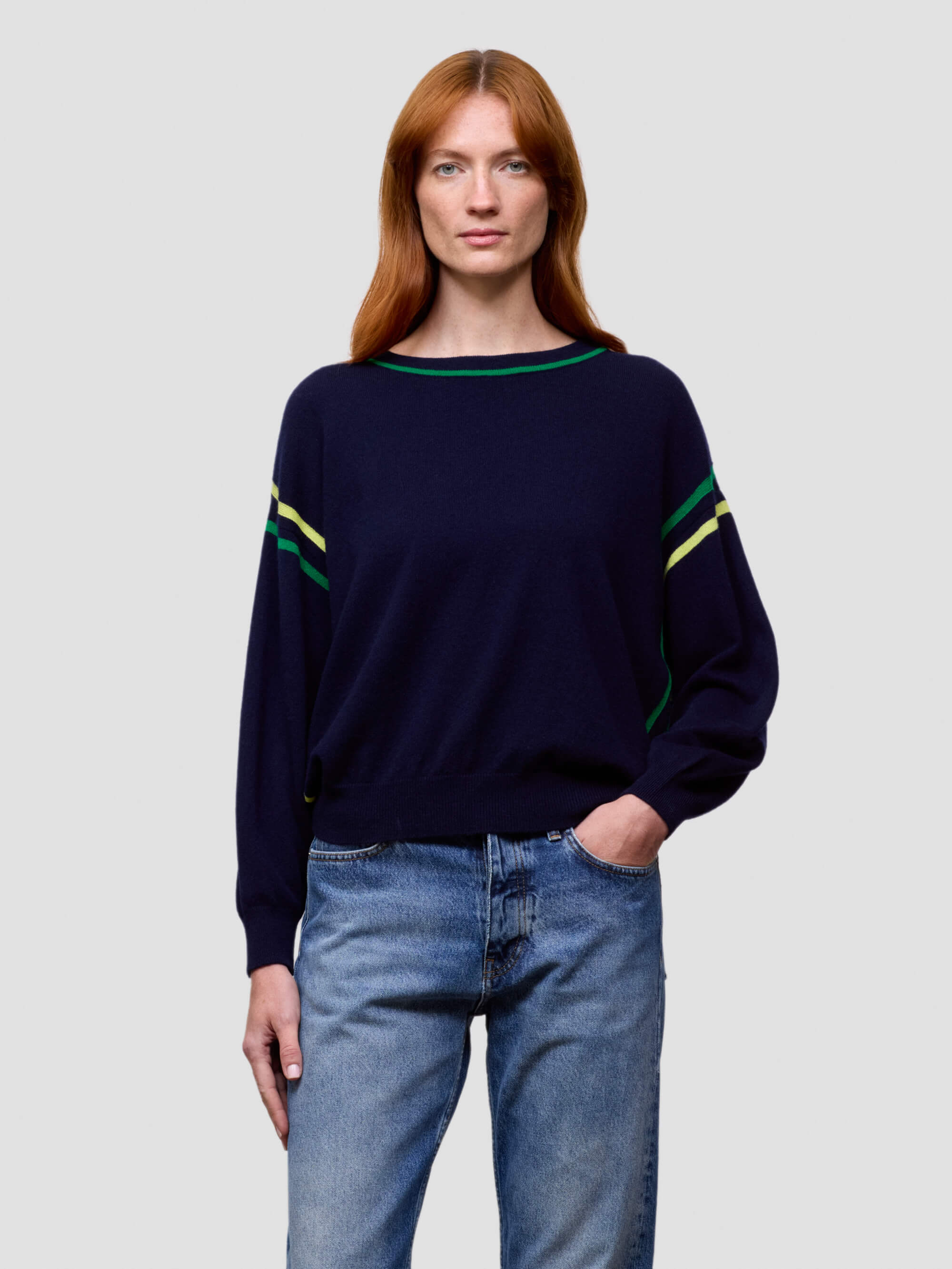 Woman wearing a navy sweater with green and yellow accents and blue jeans on a light gray background