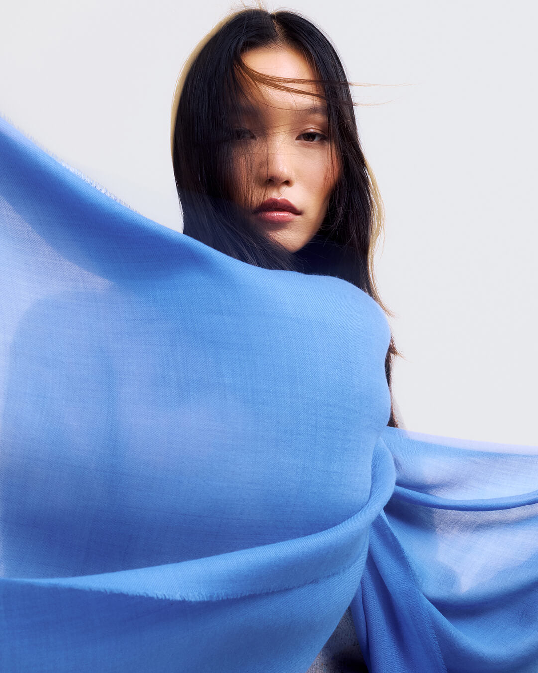 A woman with dark hair wrapped in a flowing light blue sheer cashmere scarf, set against a plain light gray background.