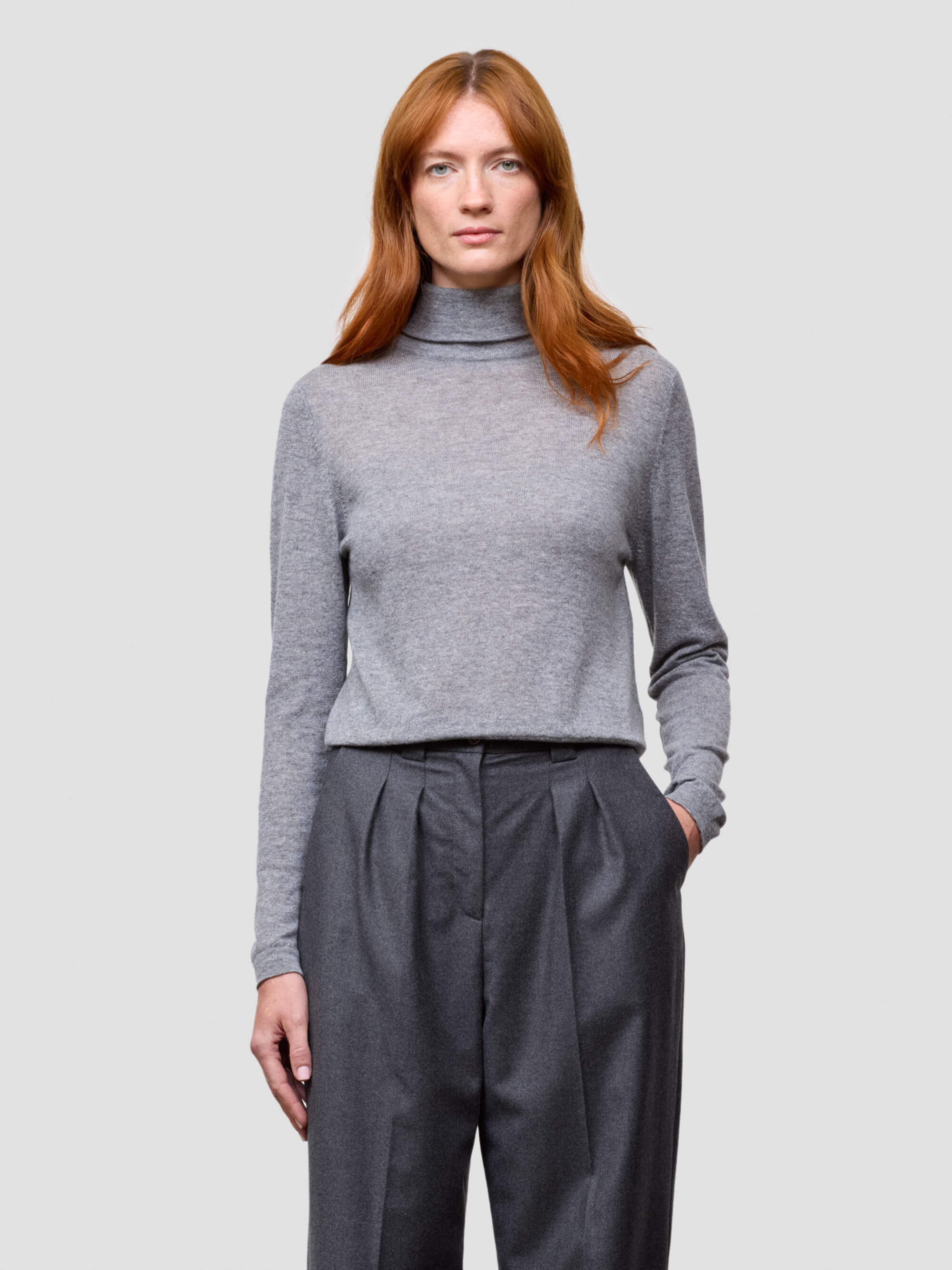 woman wearing a gray turtleneck sweater and dark gray pants on a light gray background