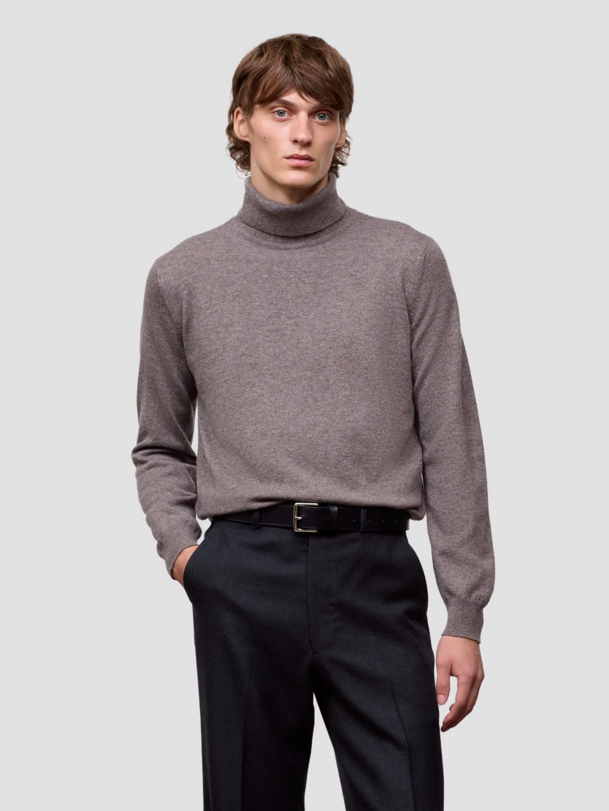Man wearing a brown turtleneck sweater and black pants on a light gray background