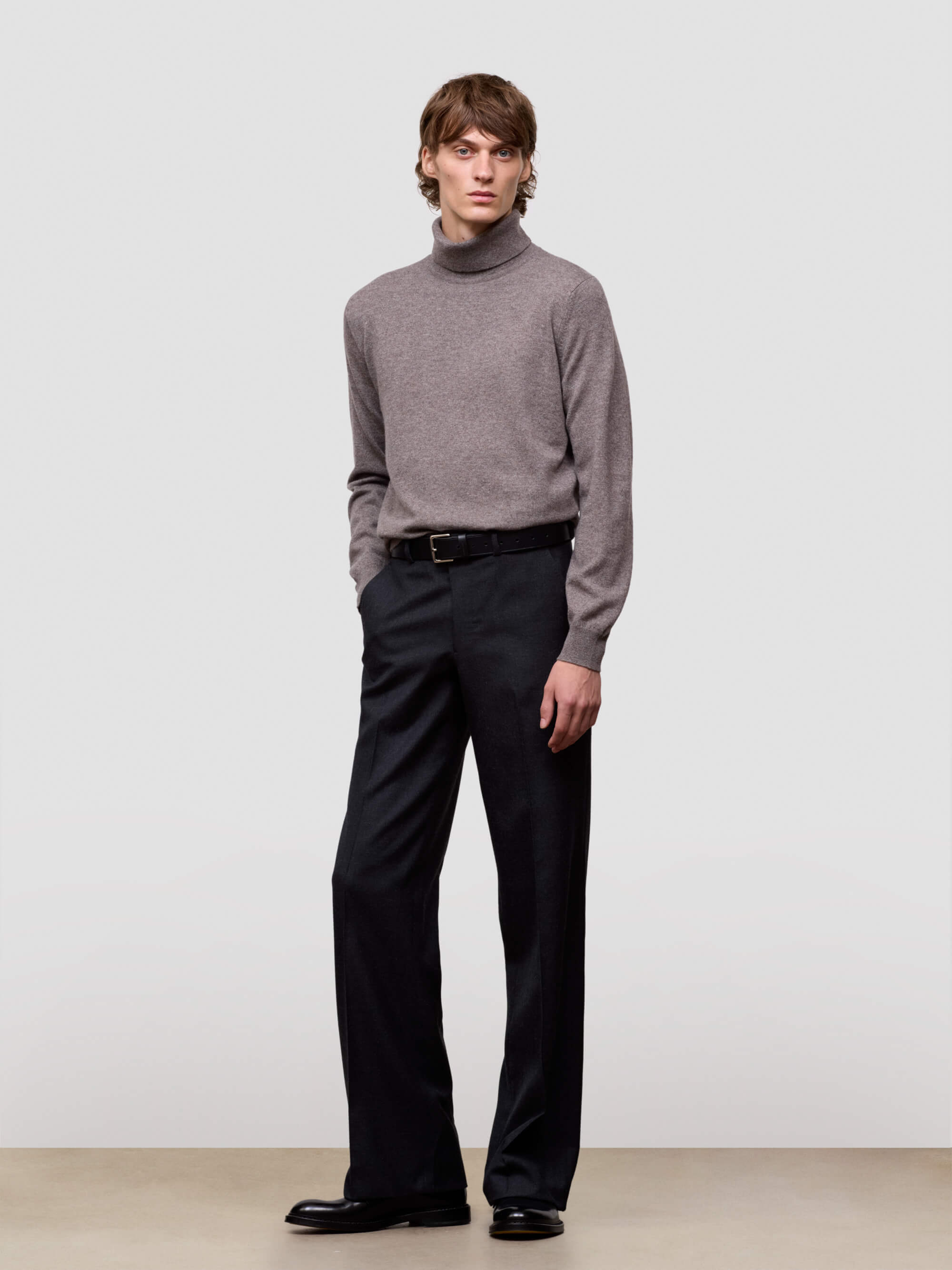 Man wearing a gray turtleneck sweater and black pants against a plain background