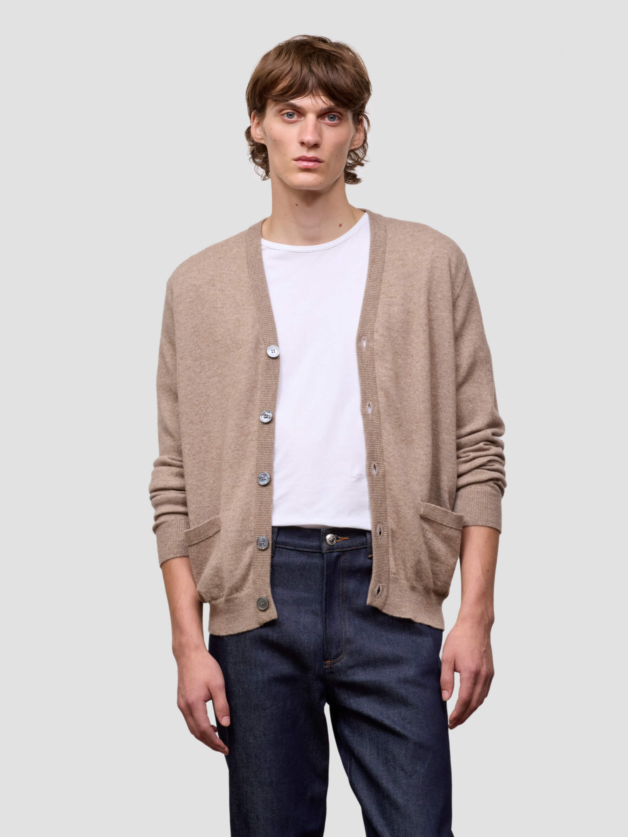 Man wearing a beige cardigan over a white shirt and dark jeans on a light gray background