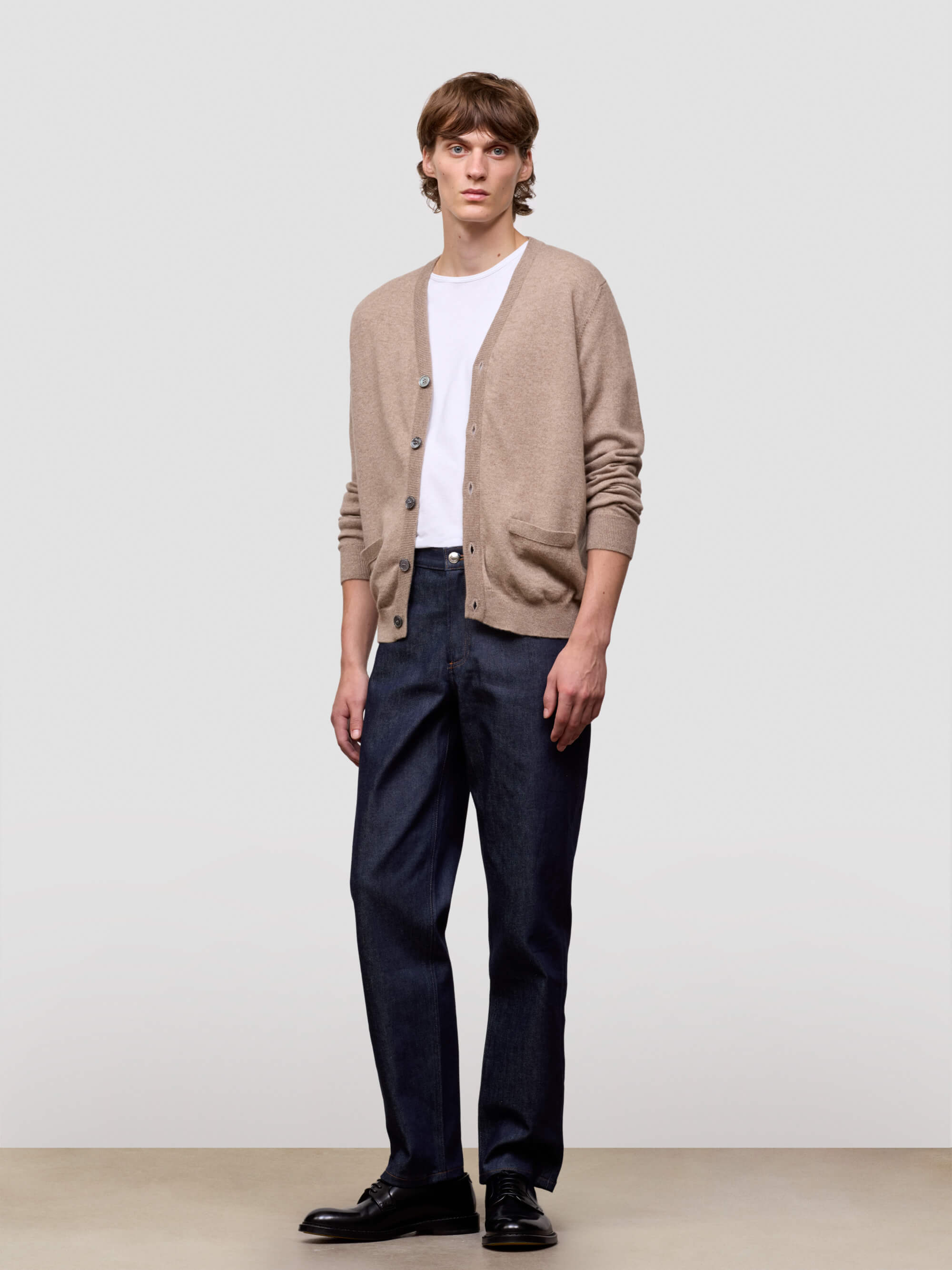 Man wearing a beige cardigan, white shirt, dark pants, and black shoes on a plain background