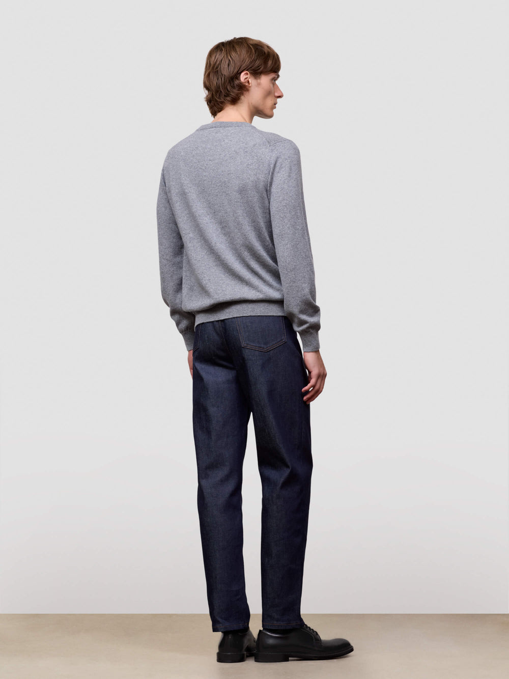 Person wearing a gray sweater and dark jeans standing against a plain white background