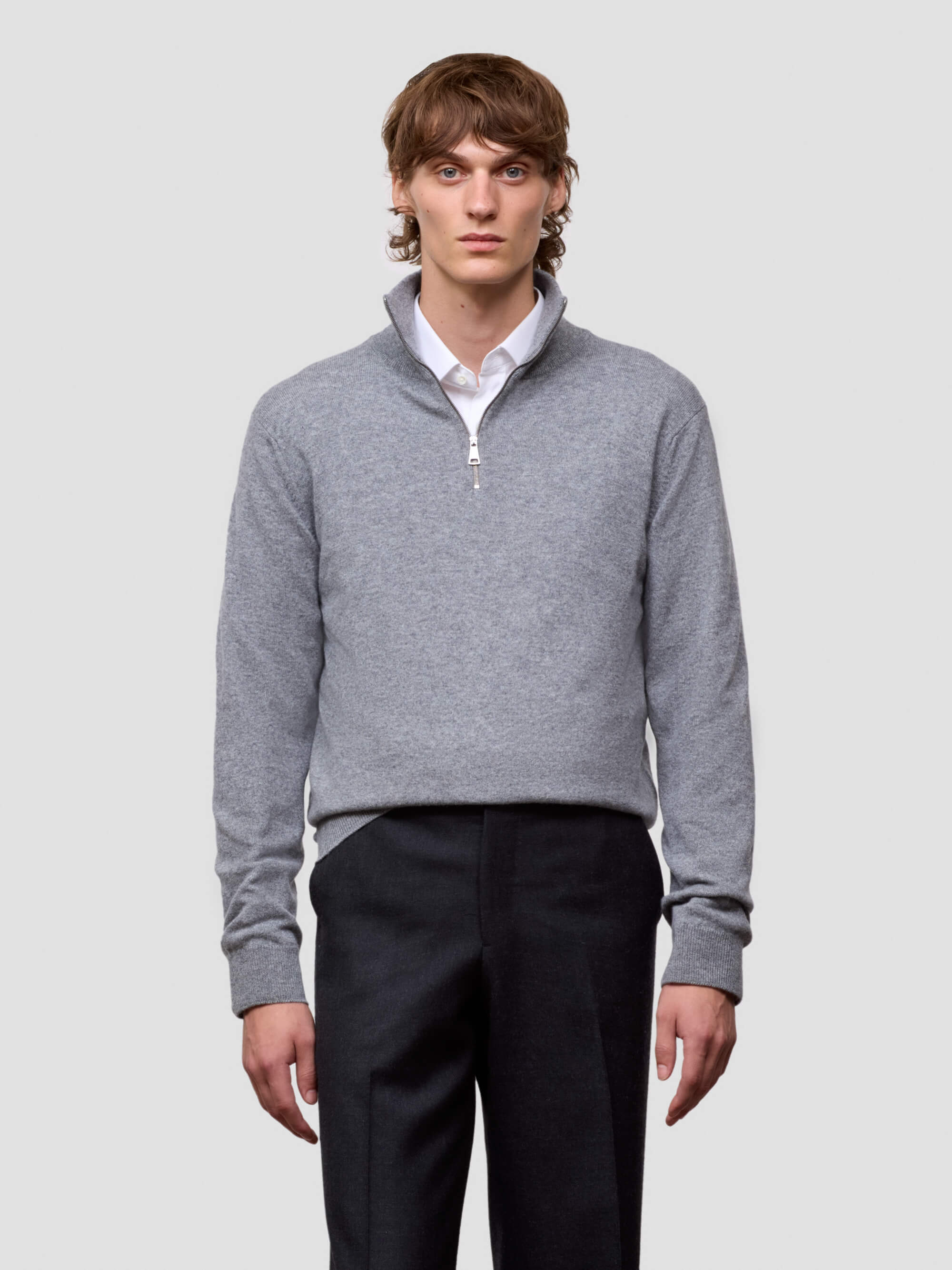 Man wearing a gray half zip sweater over a white shirt with black pants on a light gray background
