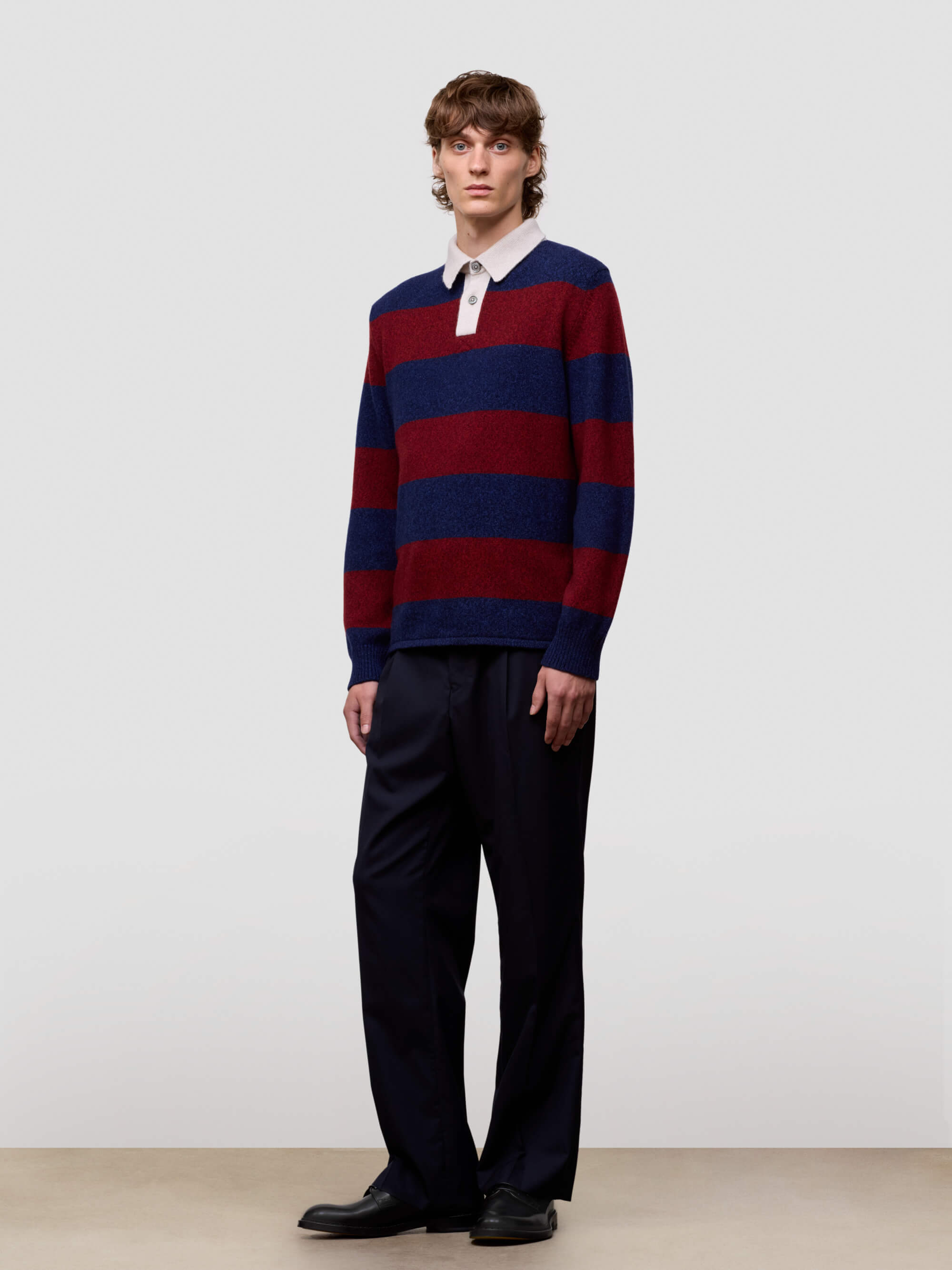 Man wearing a red and blue striped sweater with a white shirt underneath, standing against a plain background.