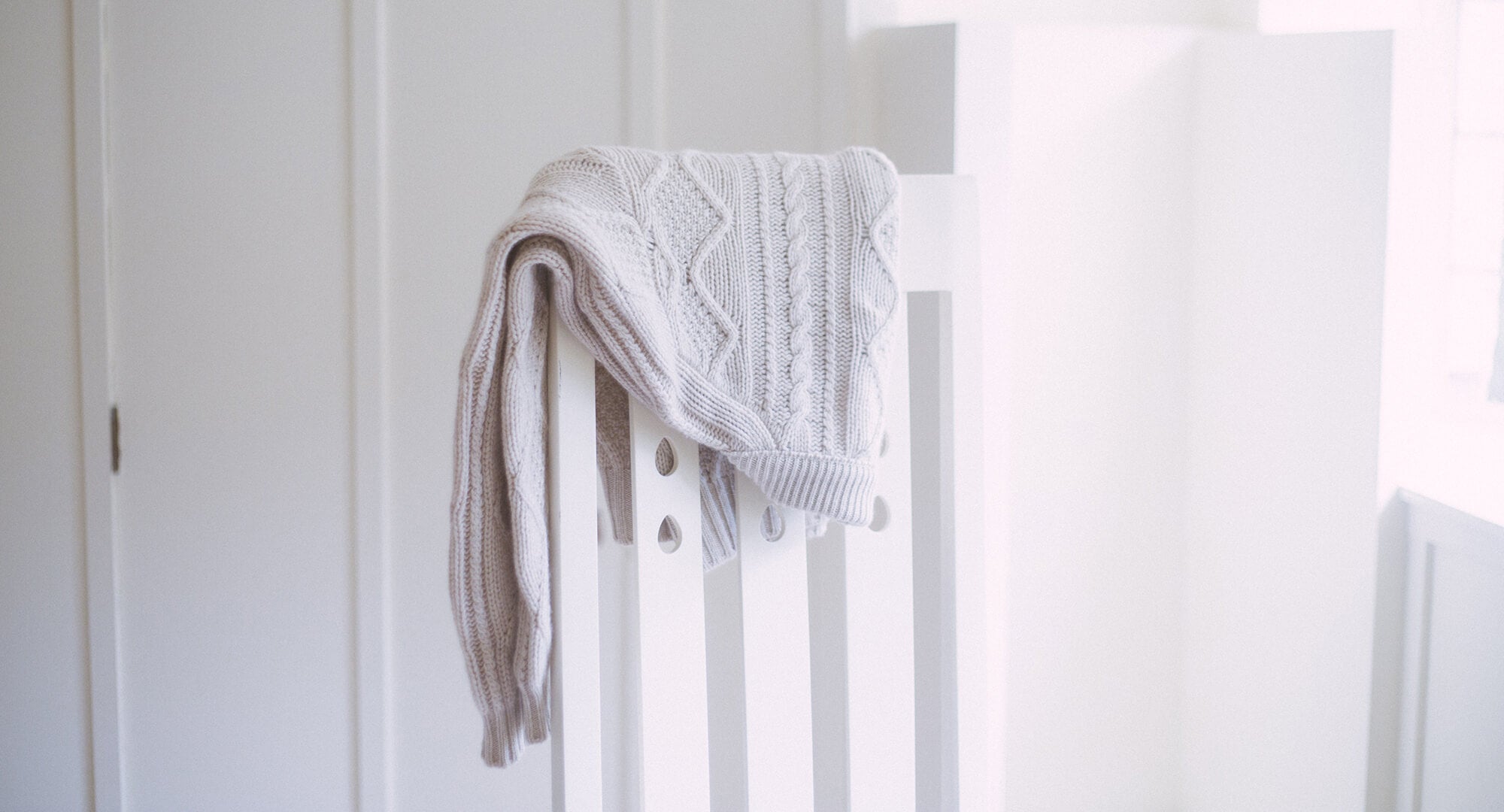 A white ribbed cashmere cardigan draped over the back of a white chair - Begg x Co.