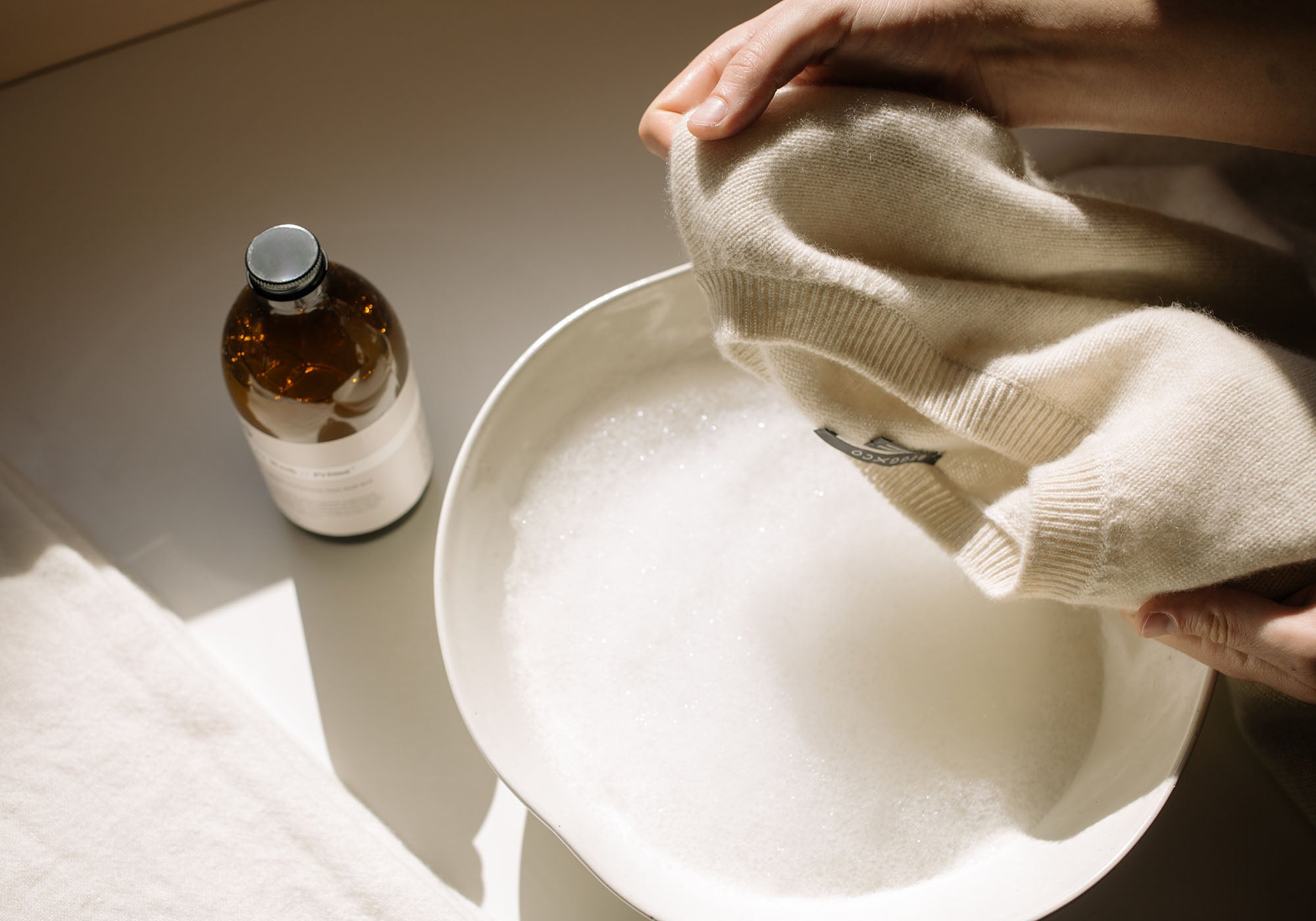 Image showing how to wash cashmere by hand, with someone ready to dip a white cashmere sweater into a ceramic bowl of bubbly water - Begg x Co 
