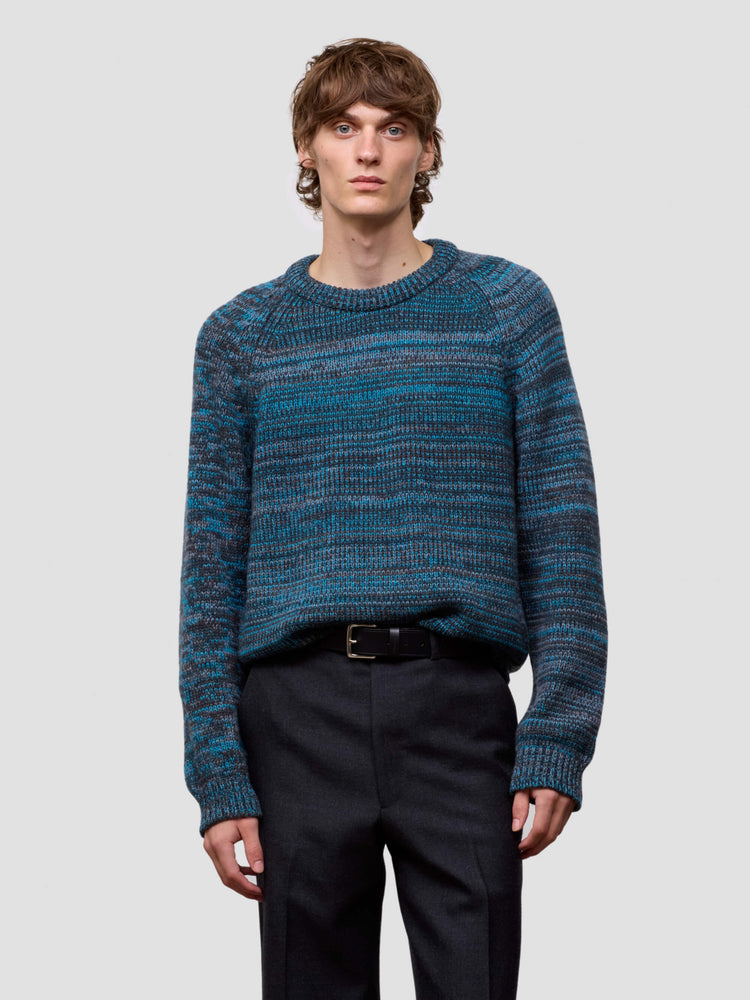 Man wearing a blue patterned cashmere sweater and black pants on a light gray background